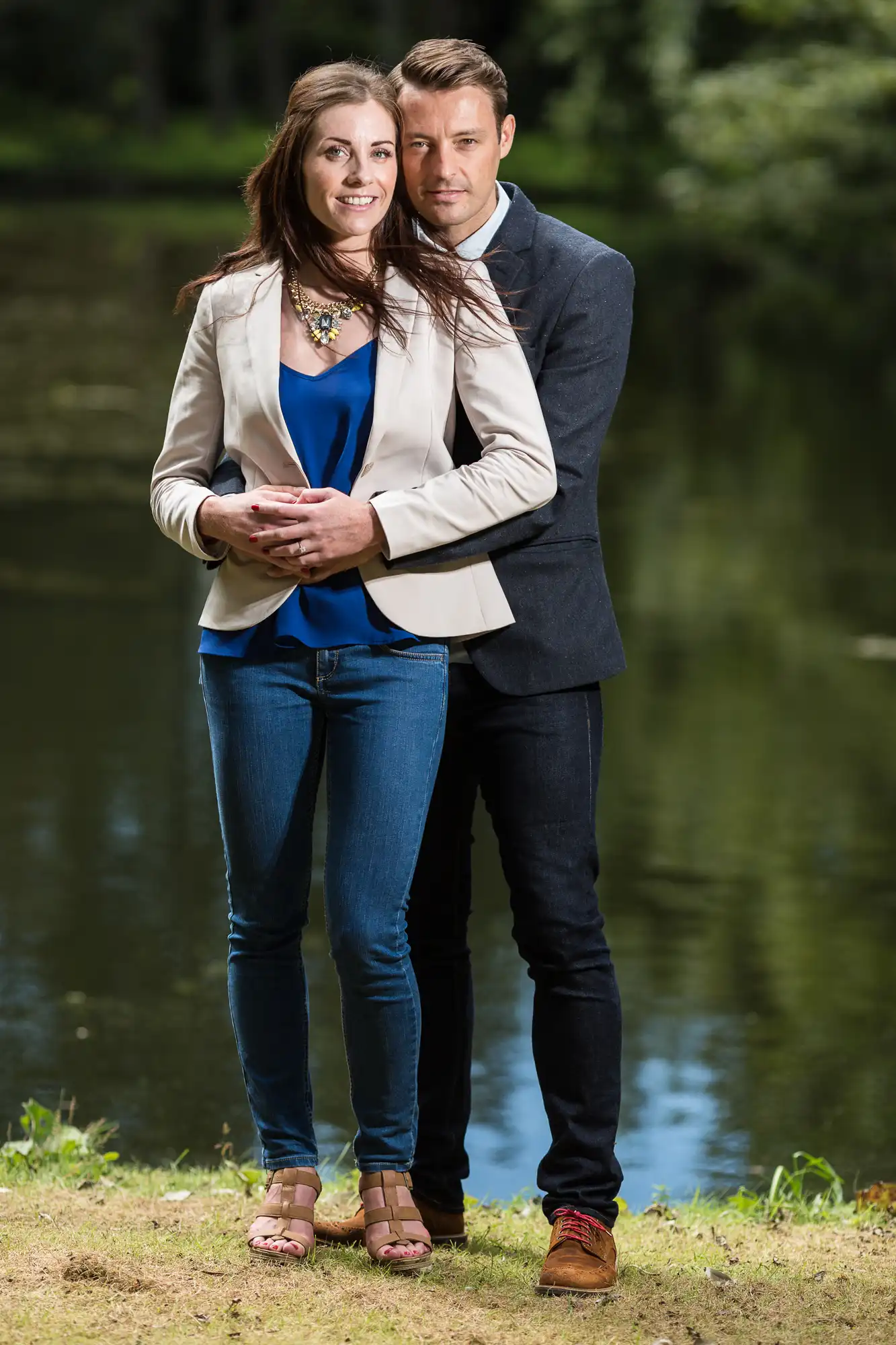 A couple standing by a lake, the woman wearing a white blazer and jeans, the man in a gray shirt and jeans, both smiling at the camera.