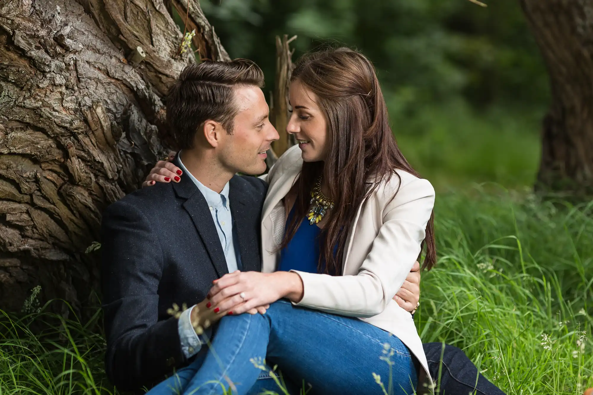 A man and a woman sitting close together by a tree, smiling and gazing into each other's eyes, surrounded by lush green grass.