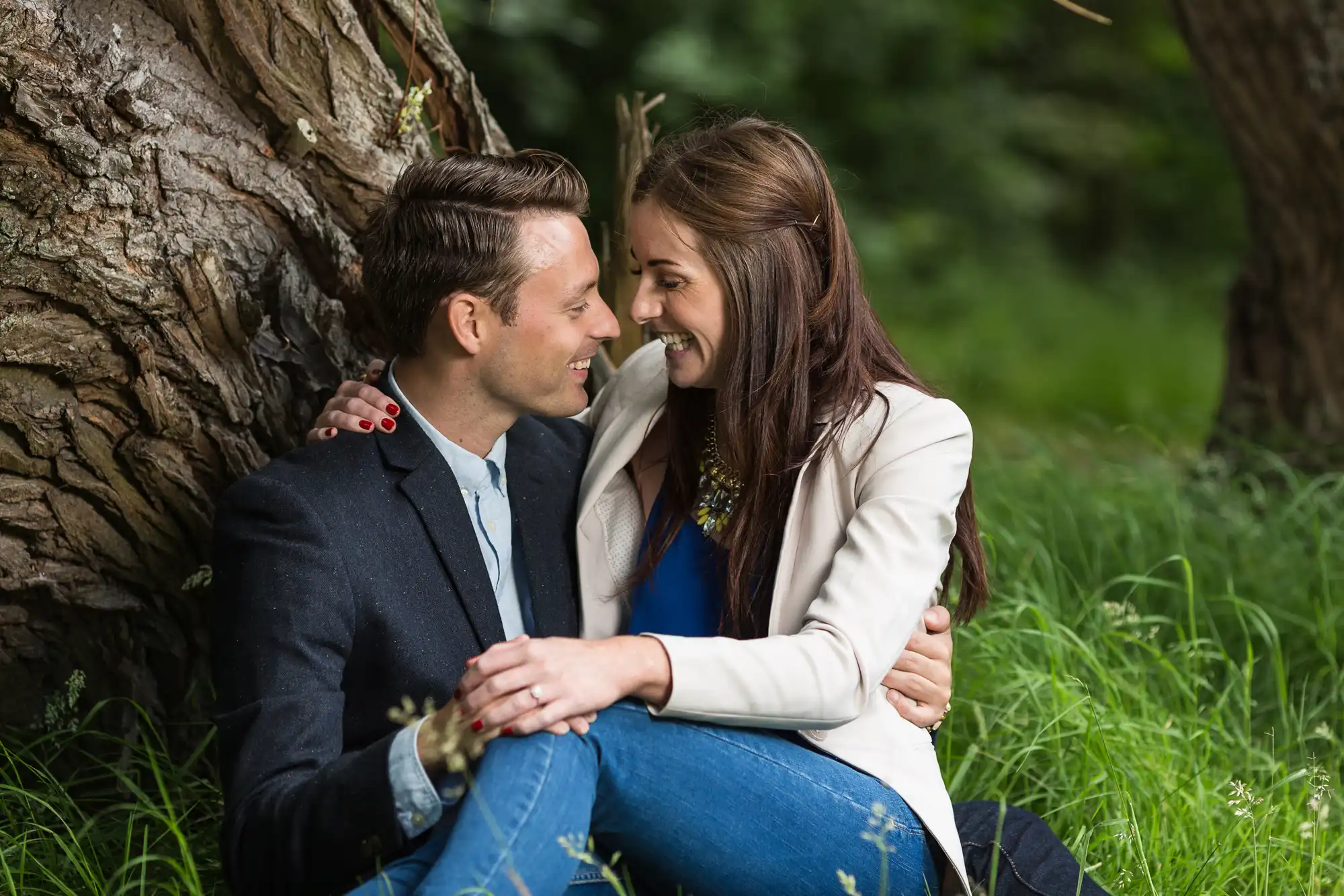 A couple sitting closely by a tree, smiling at each other in a lush green park.