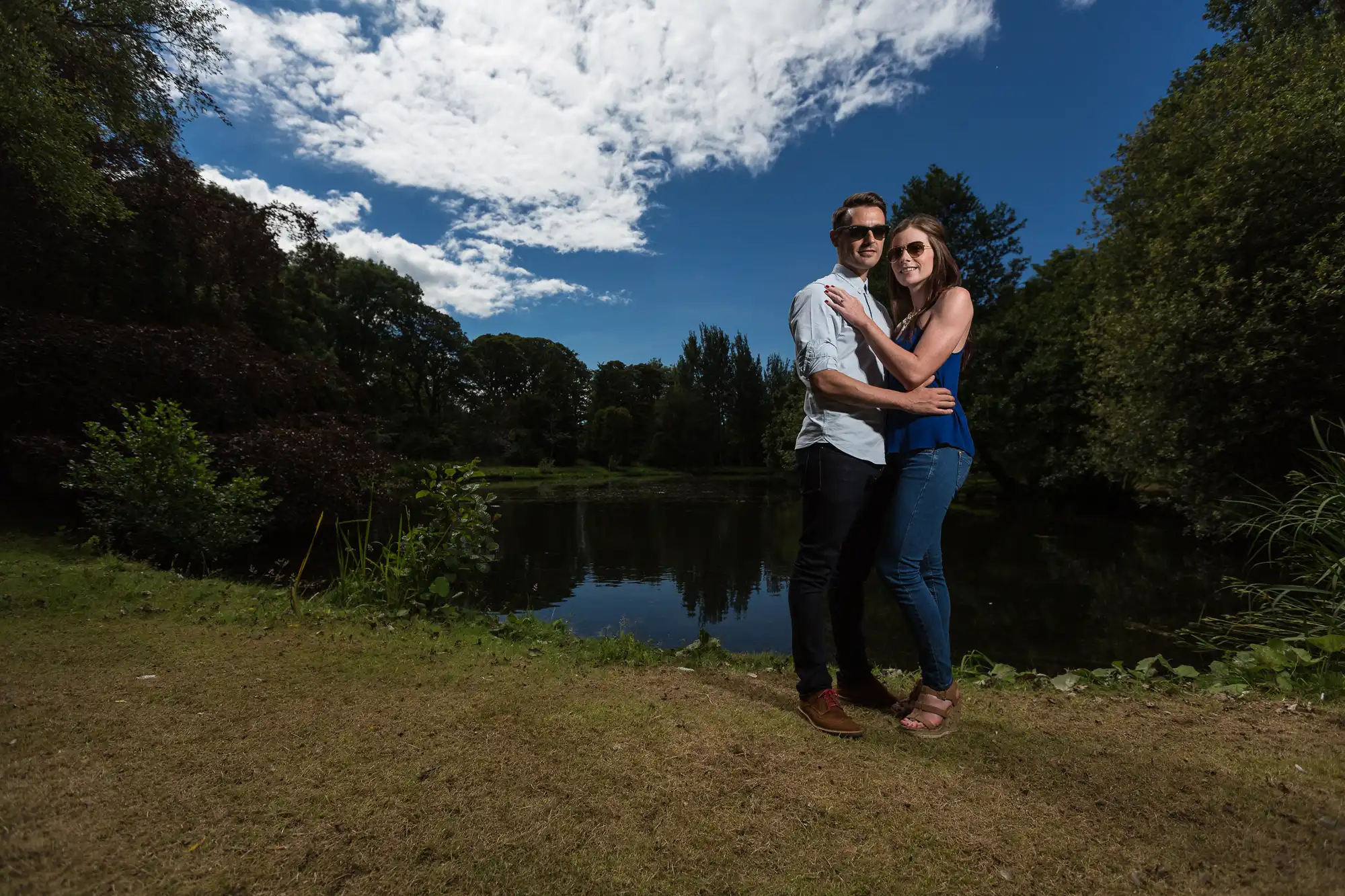A couple embracing beside a tranquil pond under a sunny sky with fluffy clouds.