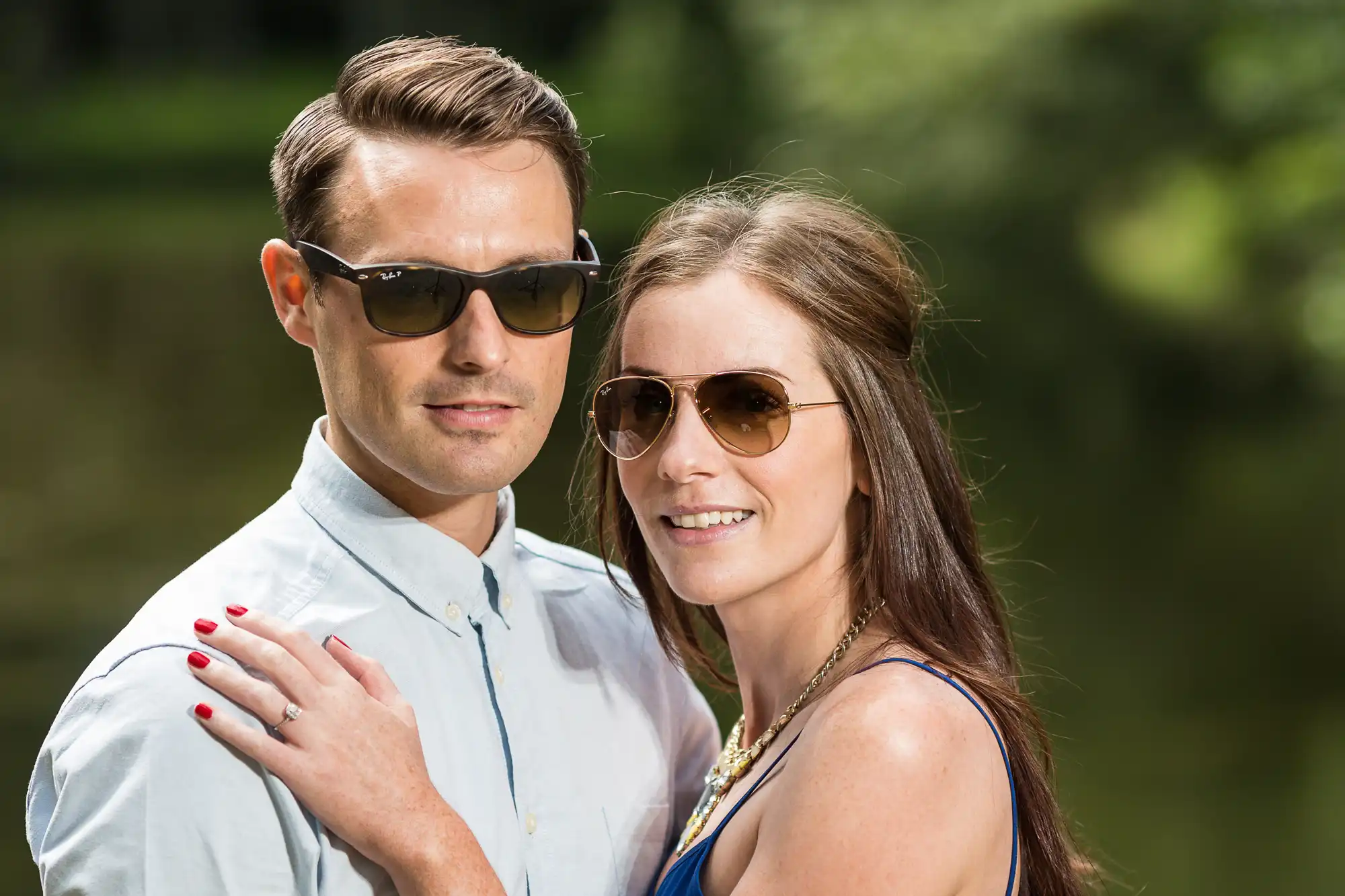 A man and woman wearing sunglasses standing close together in a park, smiling slightly towards the camera.