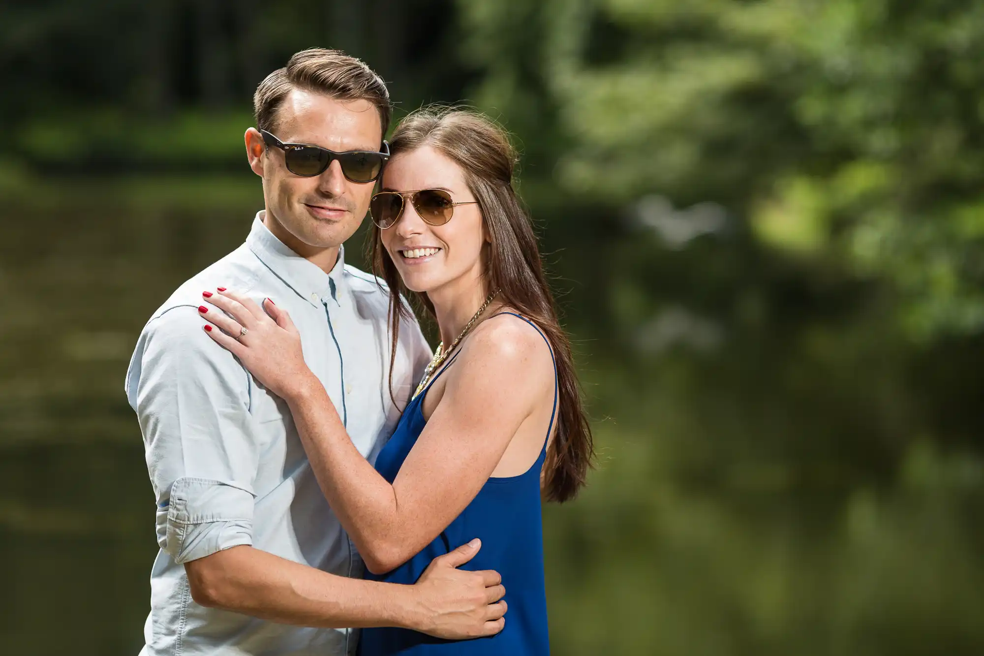 A couple embracing outdoors by a river, with the man wearing sunglasses and a white shirt, and the woman in a blue dress smiling at the camera.