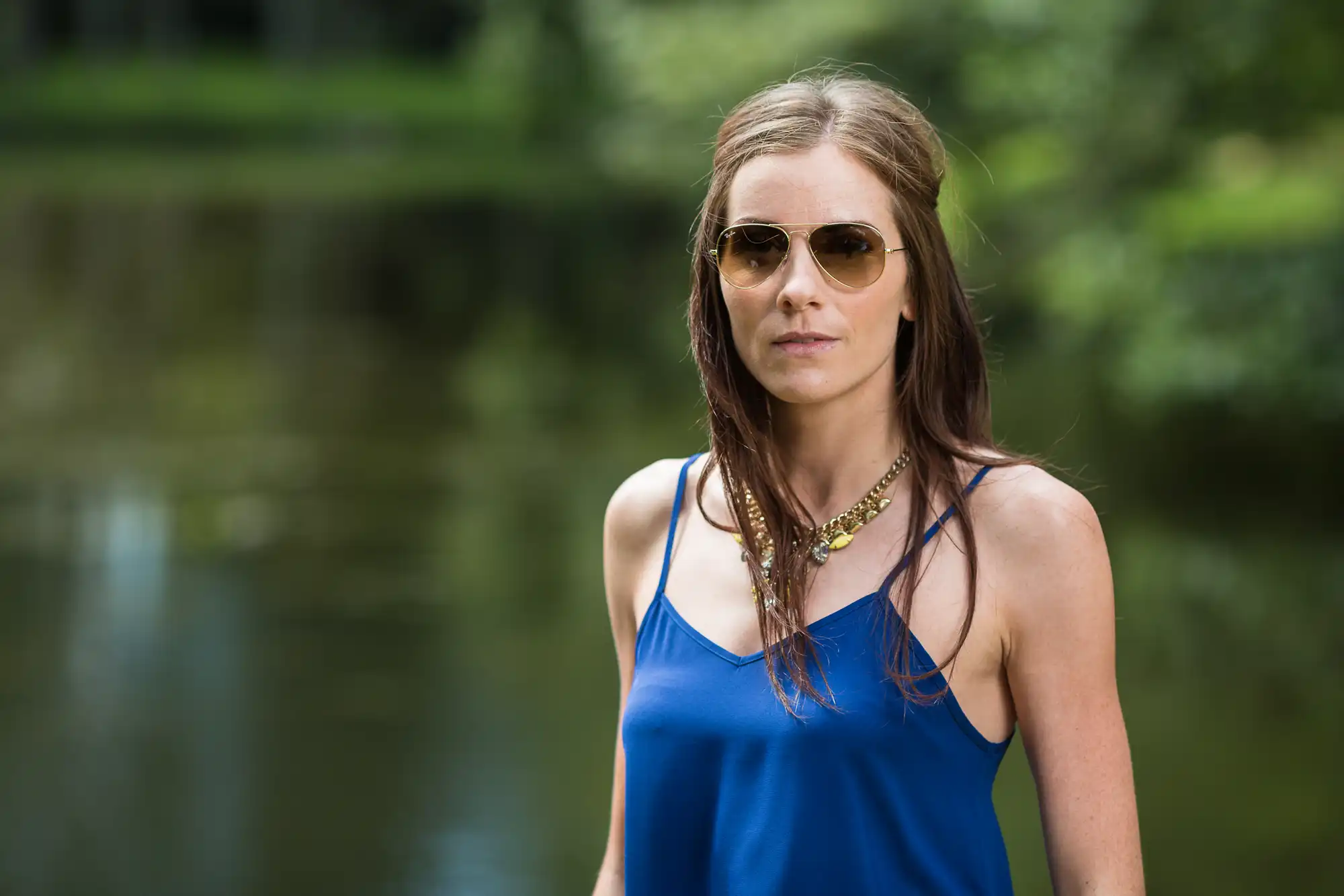 A woman wearing sunglasses and a blue top stands in front of a pond in a park.