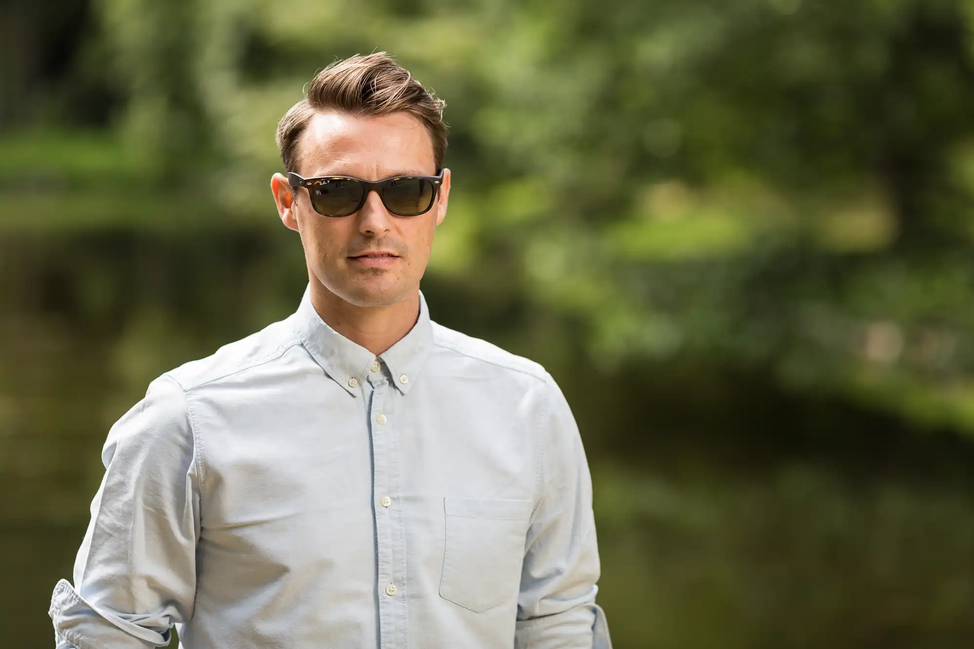 A man wearing sunglasses and a light blue shirt stands outdoors with trees and a river in the background.