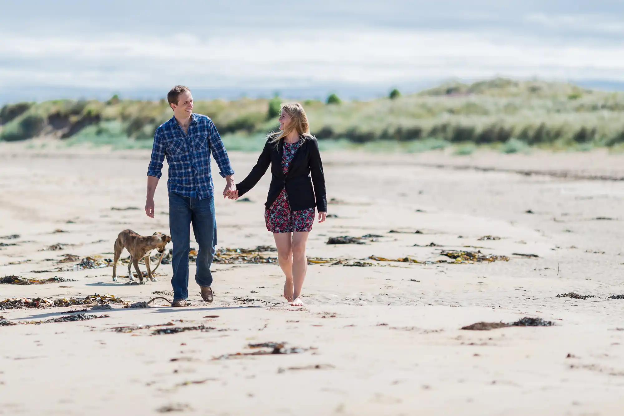 A couple walks hand-in-hand with their dog on a sandy beach, with grassy dunes in the background under a cloudy sky.