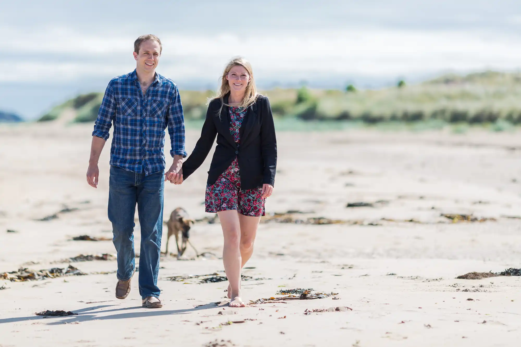 A couple walks hand-in-hand along a sandy beach with a small dog trailing behind, both smiling and casually dressed.