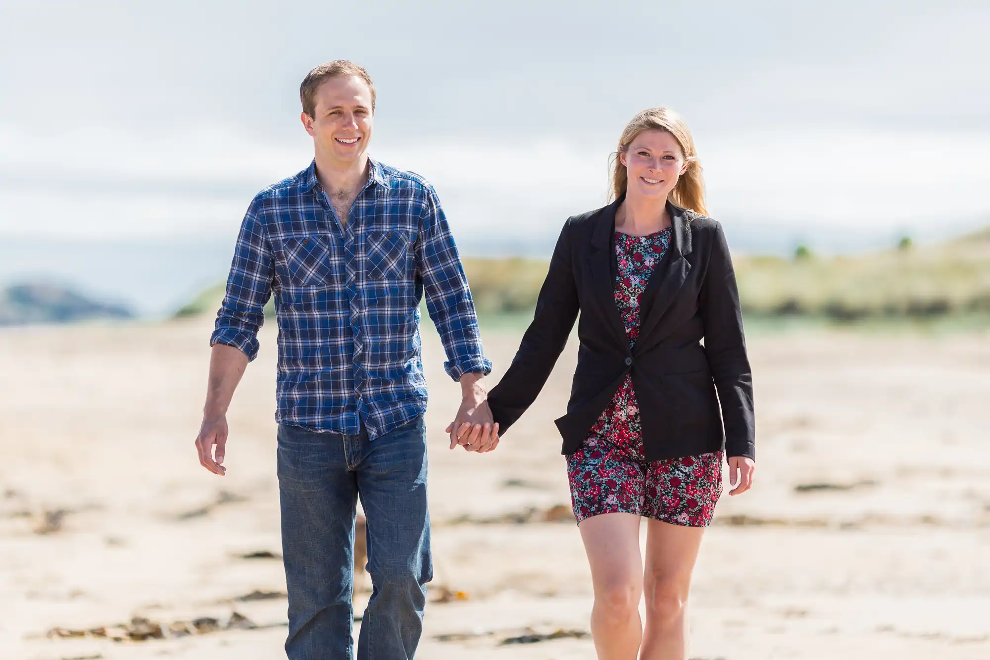 A happy couple holding hands and walking on a sandy beach, both smiling and dressed in casual outfits.