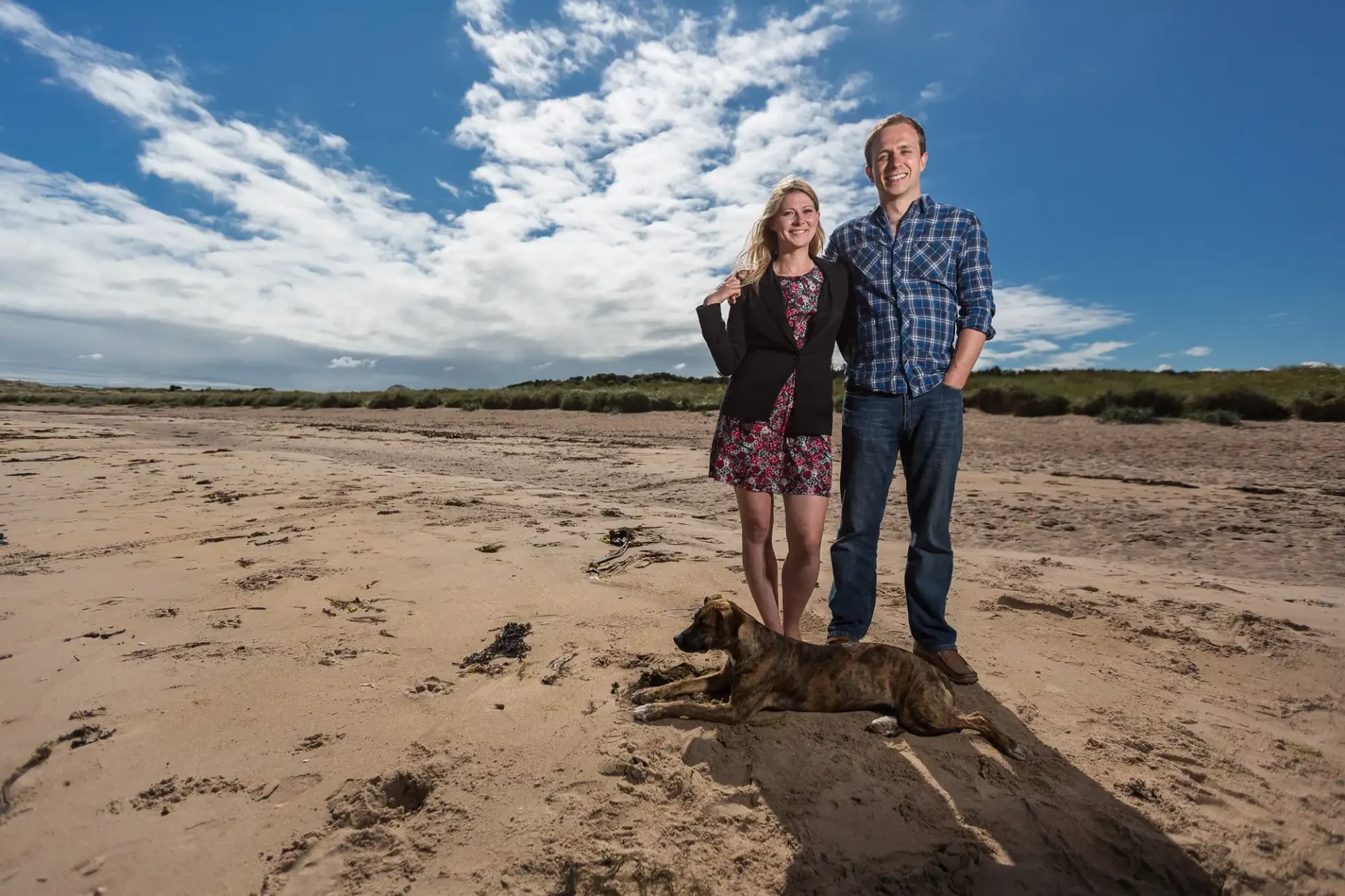 Yellowcraig Beach engagement photographer image of a woman and a man stand together on a sandy beach under a blue sky with clouds, while a brown dog lies on the sand in front of them.