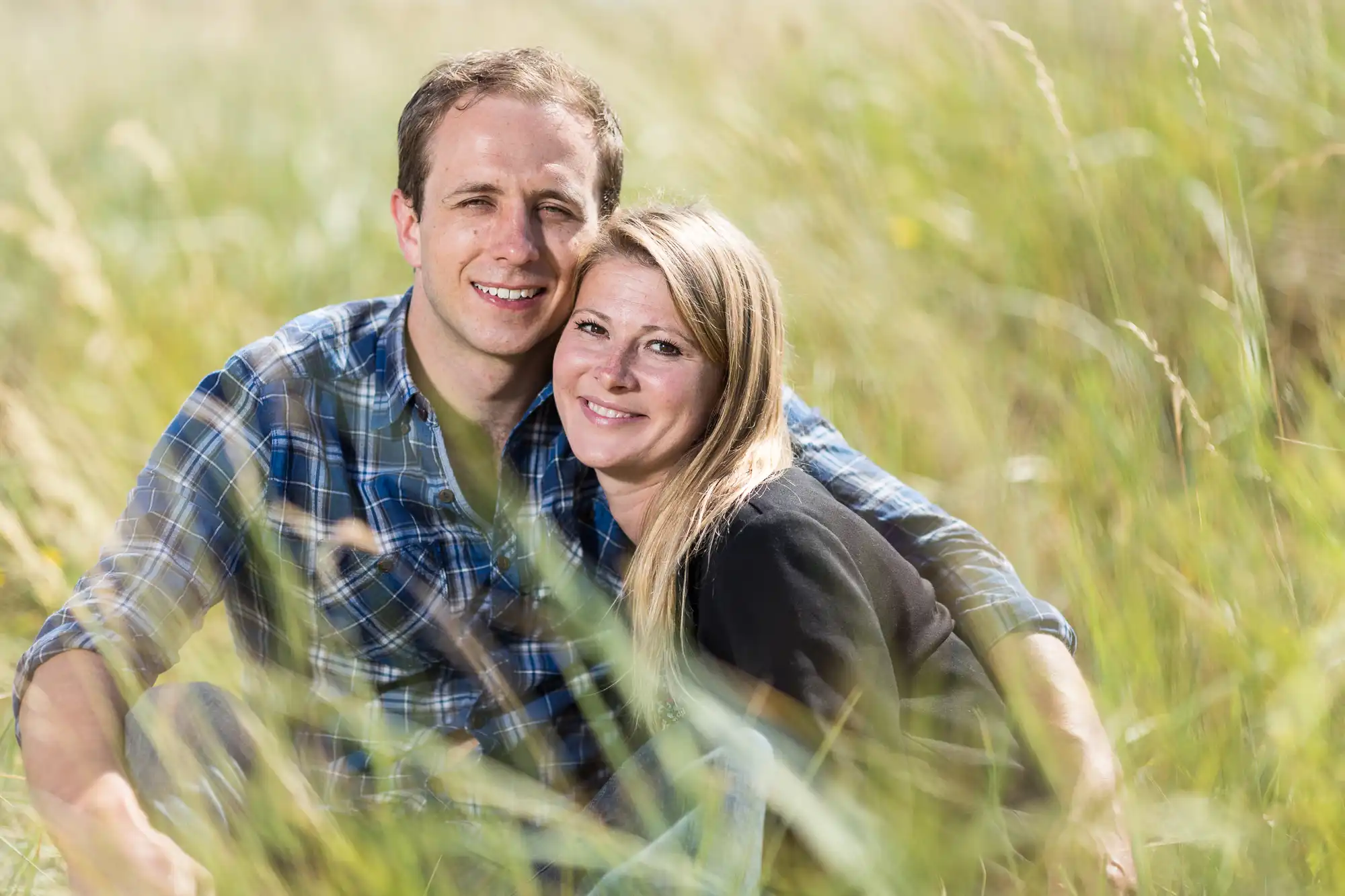 A couple smiling and embracing in a sunny meadow, surrounded by tall grass.