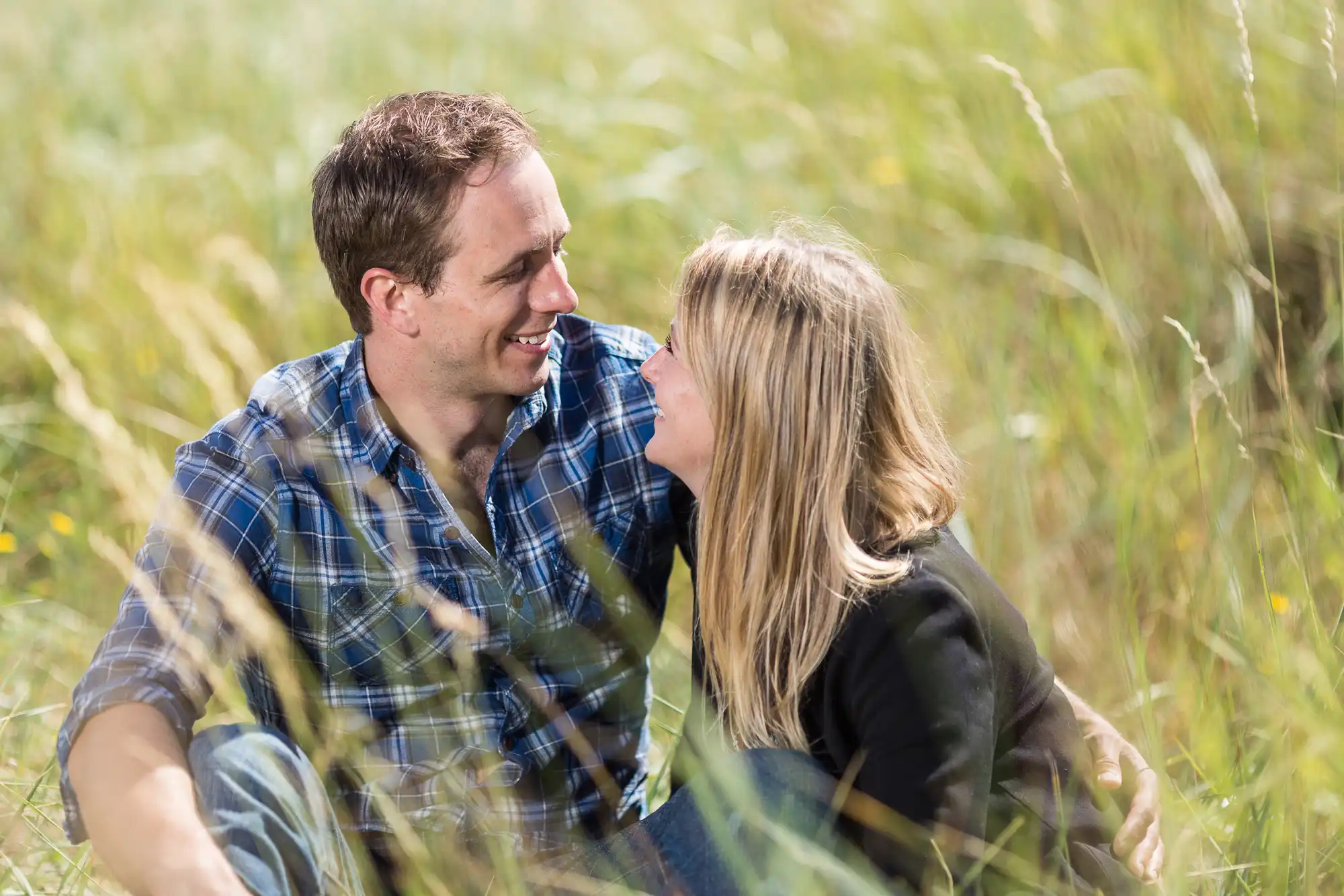 A couple sits closely in a field, smiling at each other among tall grasses, with a soft focus on surrounding greenery.