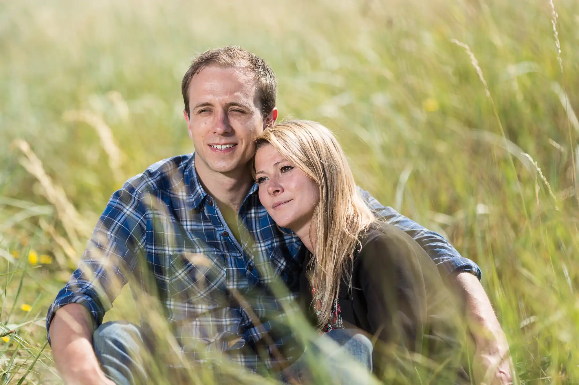 A couple sitting closely together in a sunny meadow, smiling gently and looking into the distance.