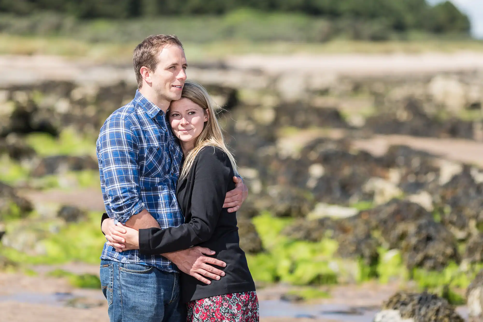 A couple embracing on a rocky beach, smiling gently, with blurred greenery and ocean in the background.