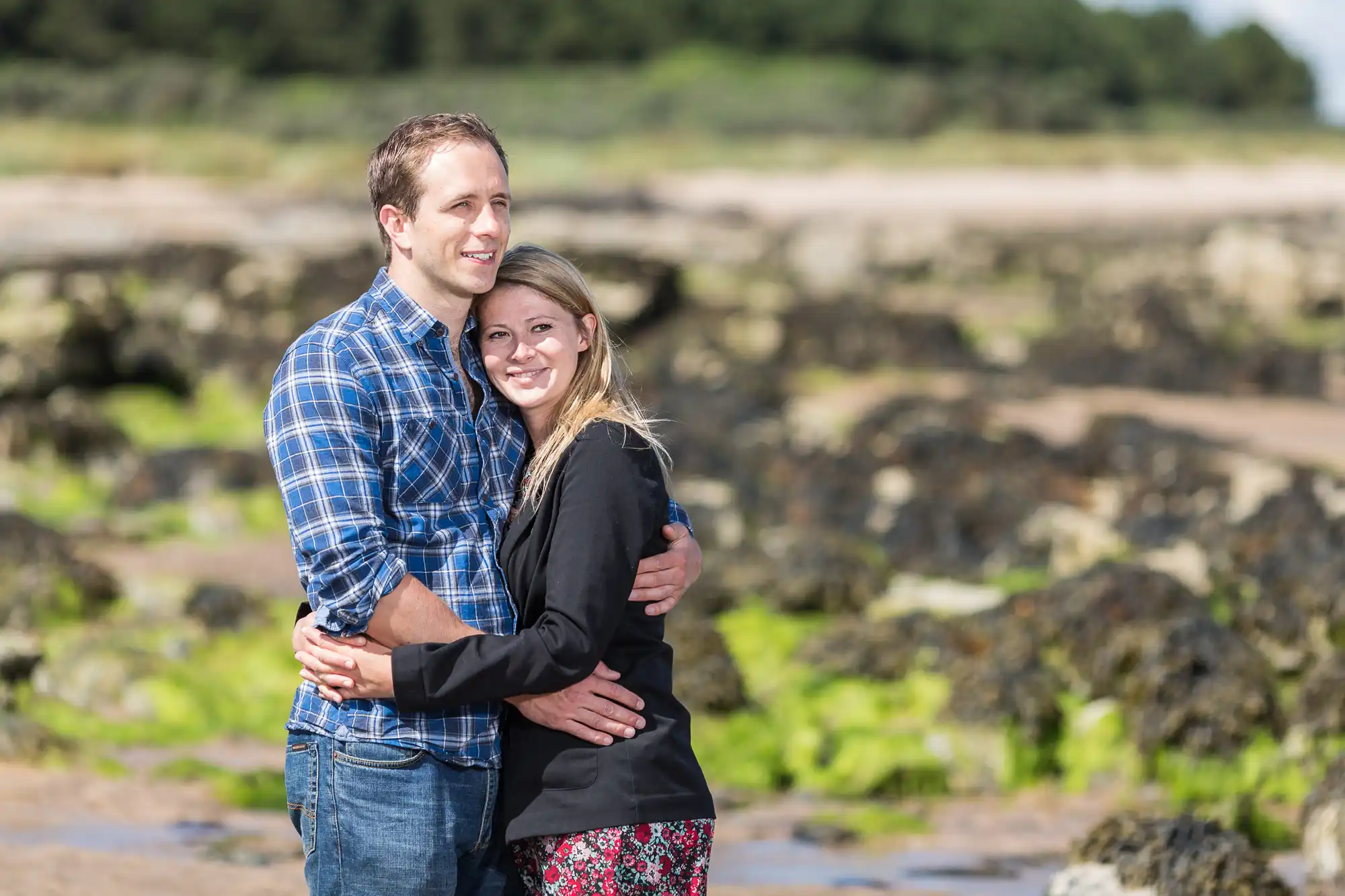 A couple embracing on a rocky beach, smiling towards the camera, with a clear sky and coastal terrain in the background.