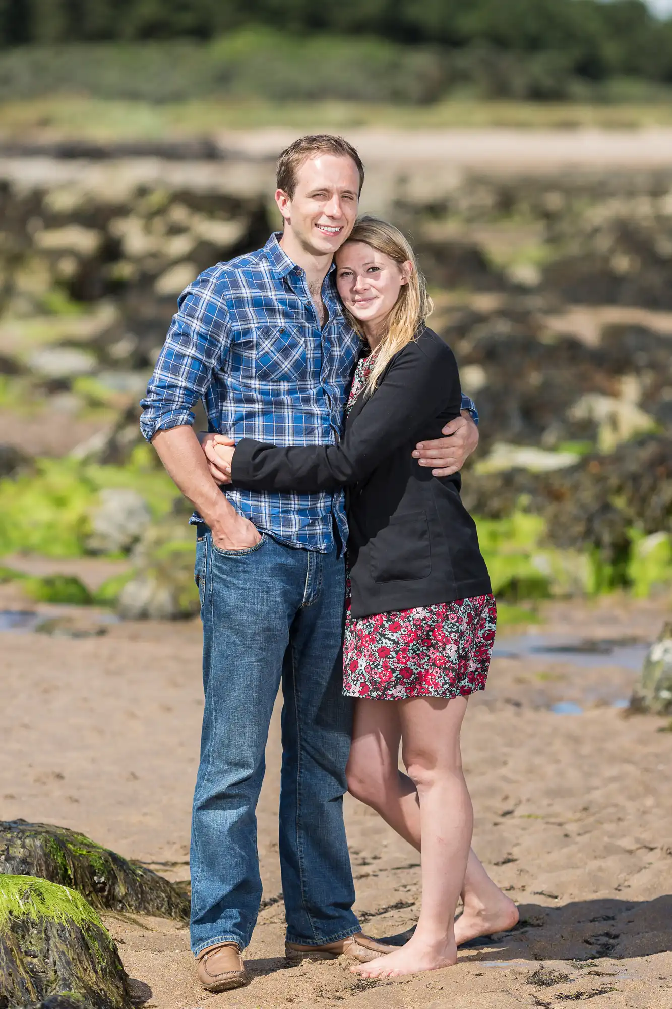 A couple embracing on a sandy beach, smiling at the camera, with ocean and rocks in the background. the man is wearing a plaid shirt and jeans; the woman is in a black jacket and floral skirt.
