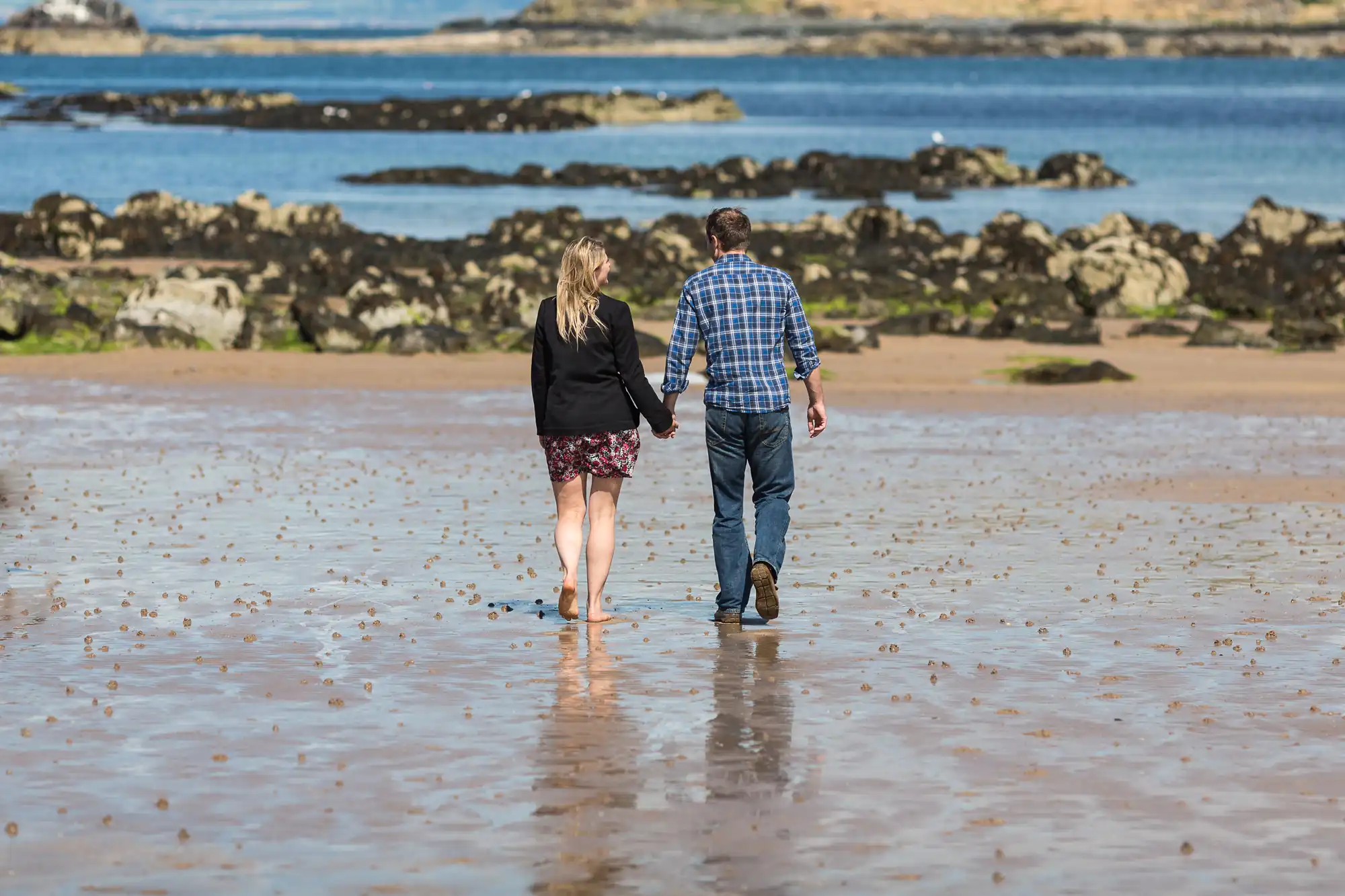 A couple walks hand in hand on a wet sandy beach, with rocks and the ocean in the background.