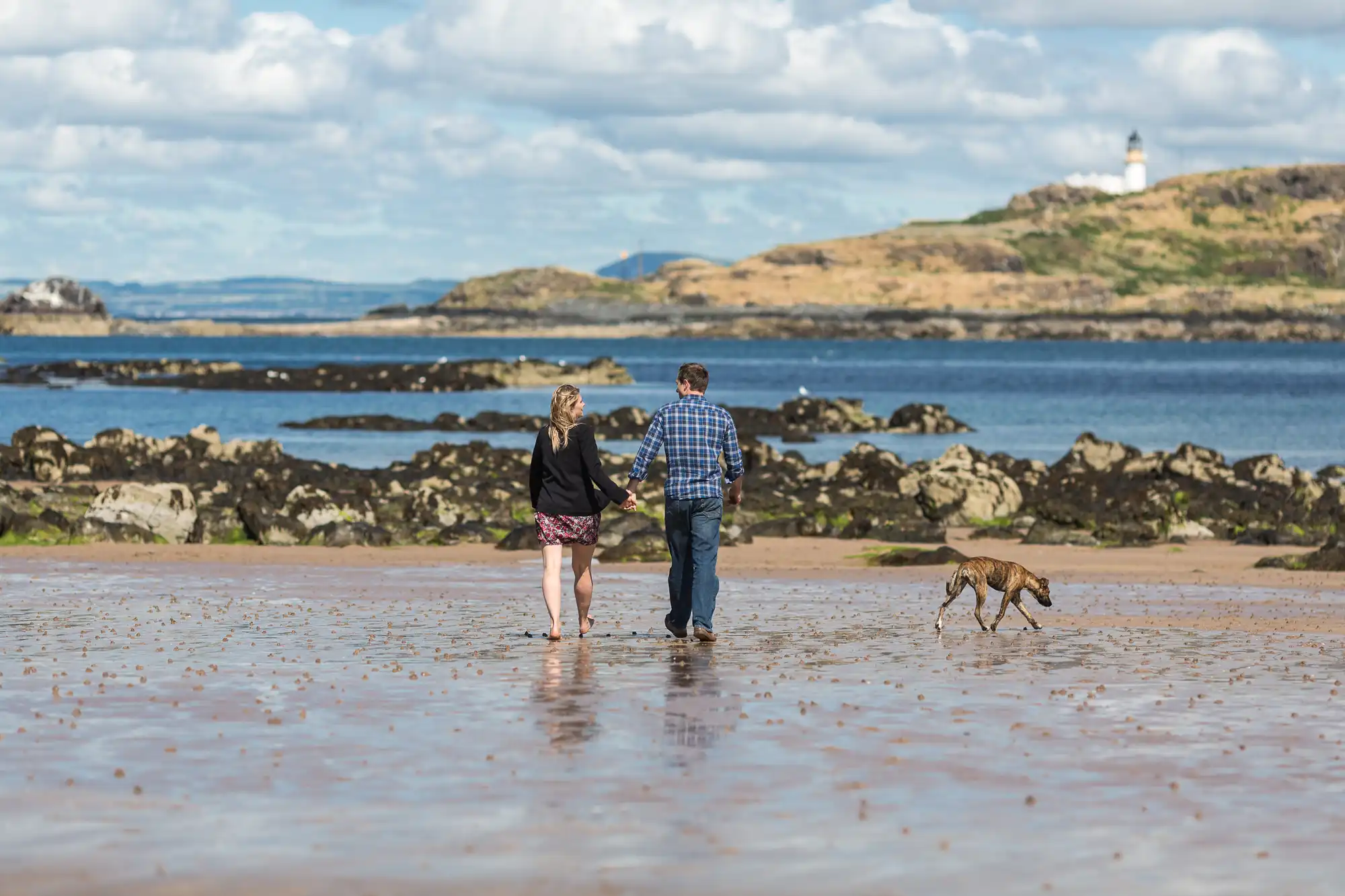 A couple holding hands walks a dog along a wet sandy beach with a lighthouse and rocky islets in the background under a cloudy sky.