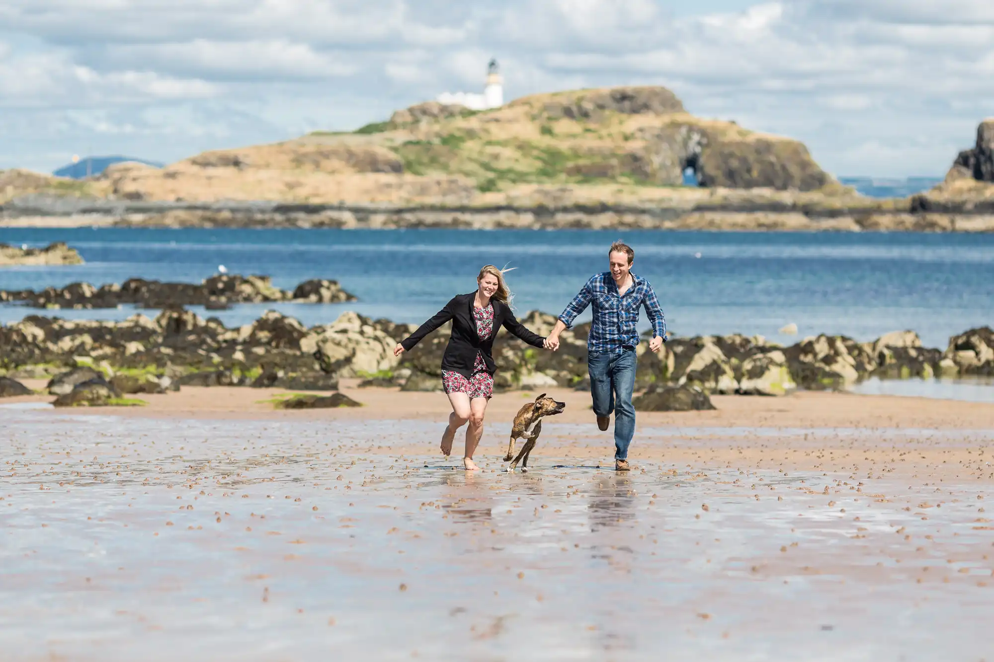Two people and a dog running on a sandy beach with rocks and a lighthouse in the background.
