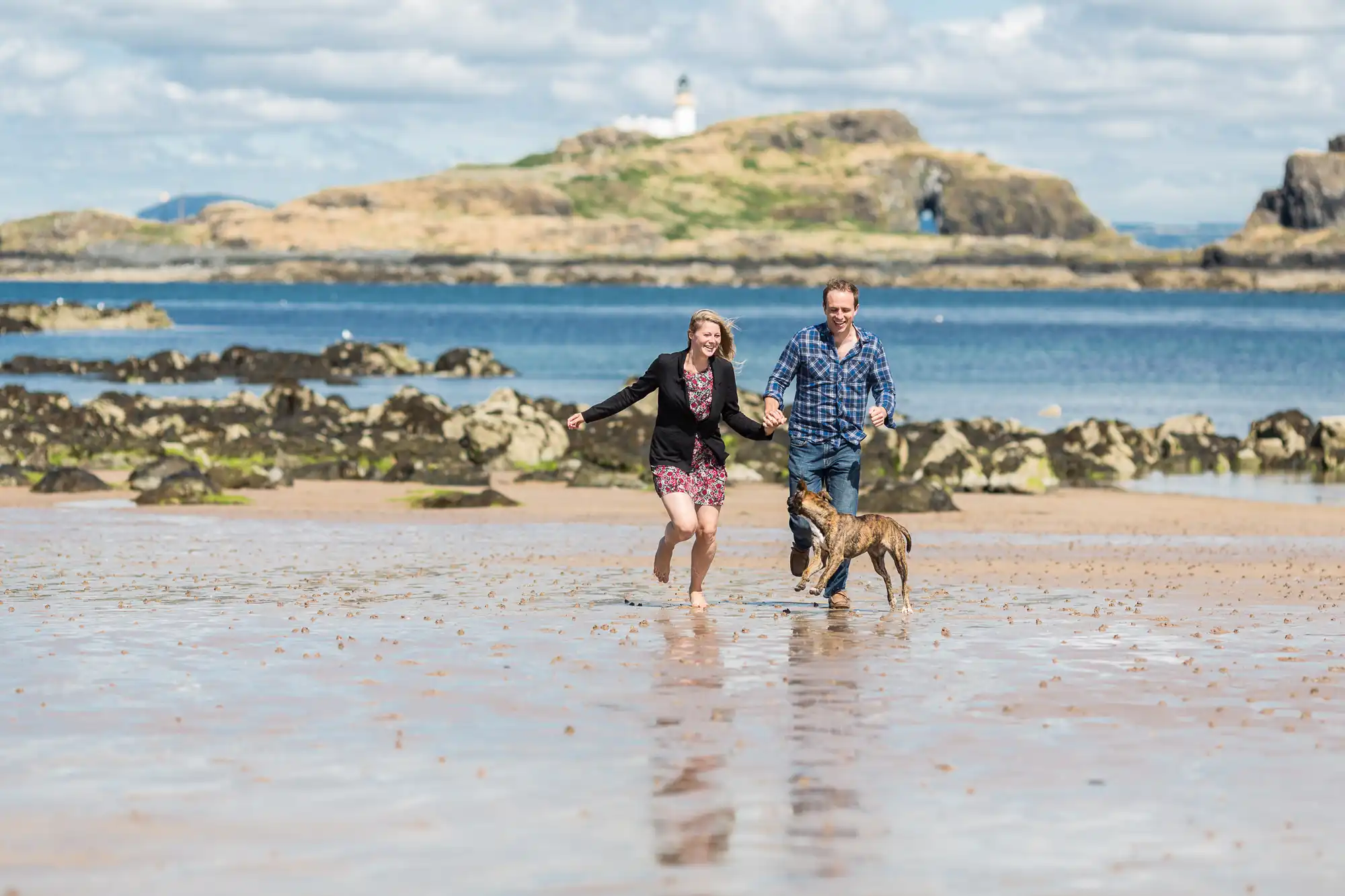 A couple runs joyfully on a sandy beach with their dog, with a lighthouse on a distant island and blue sky with clouds in the background.