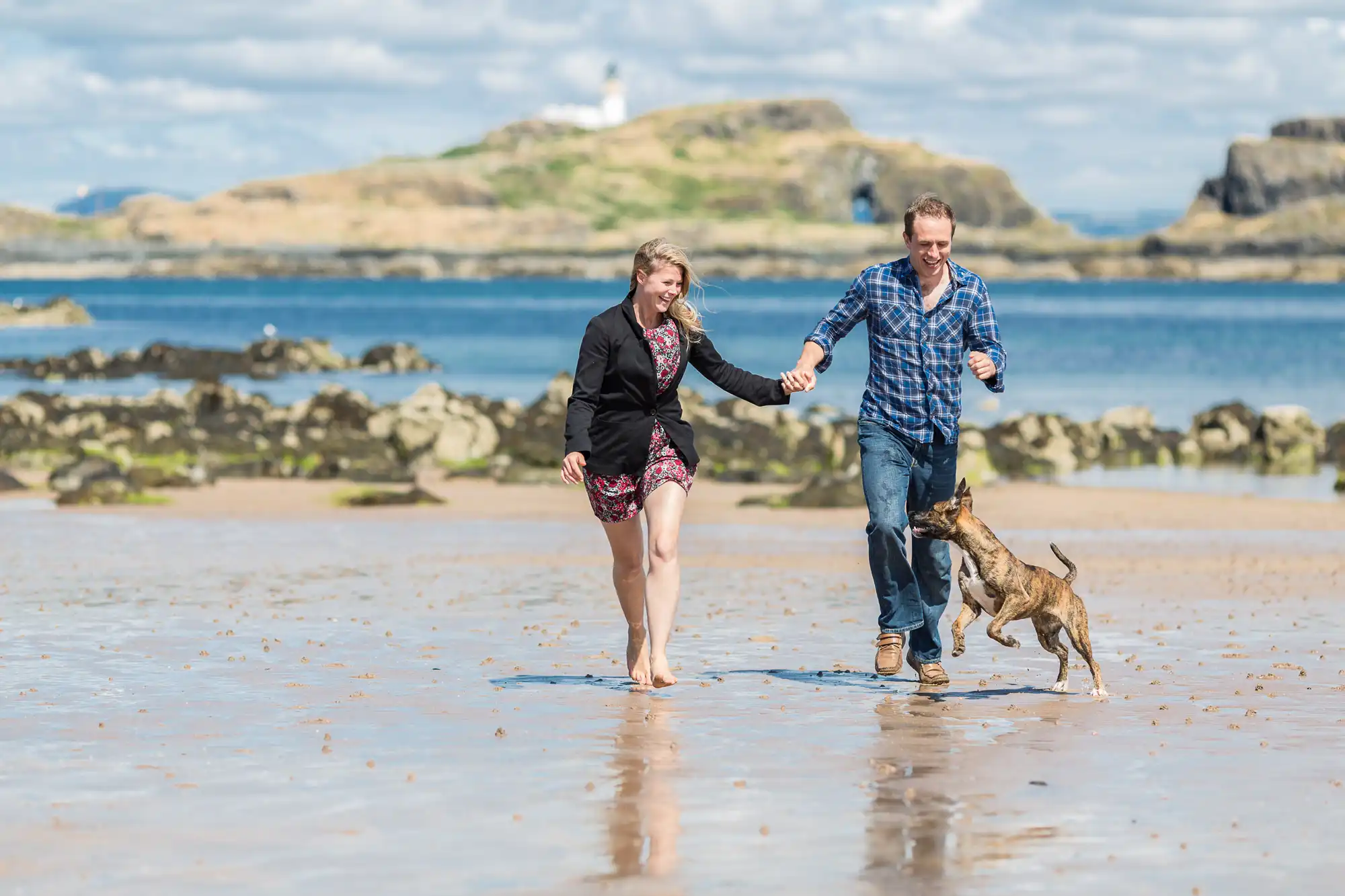 A couple walking with their dog on a sandy beach, holding hands and smiling, with a lighthouse and blue sky in the background.
