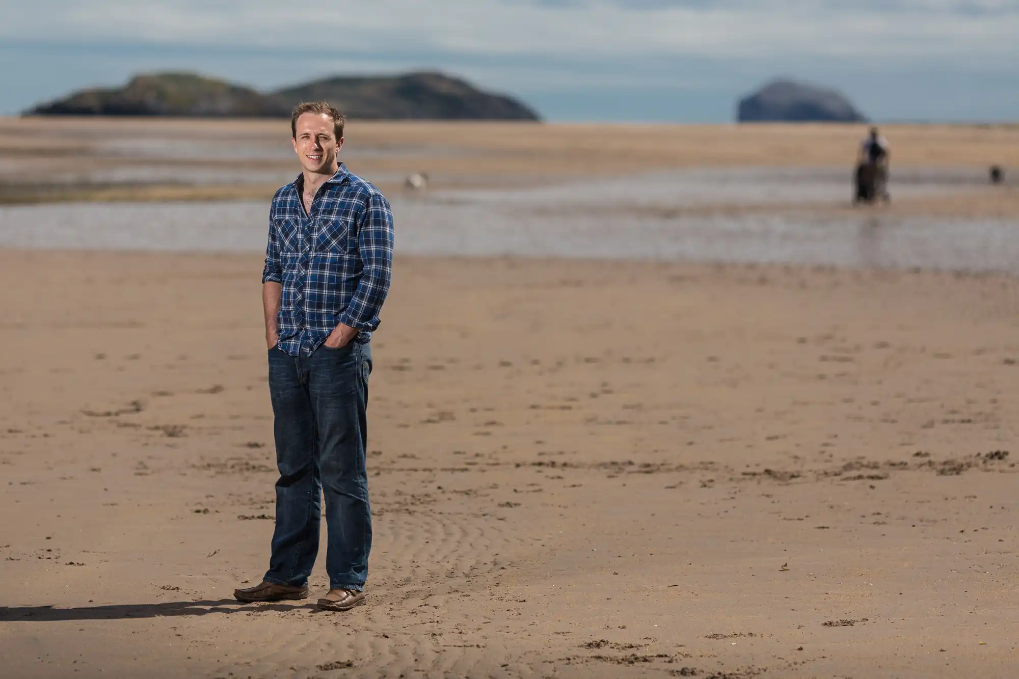 Man in a blue plaid shirt and jeans smiling on a sandy beach with distant islands in the background.
