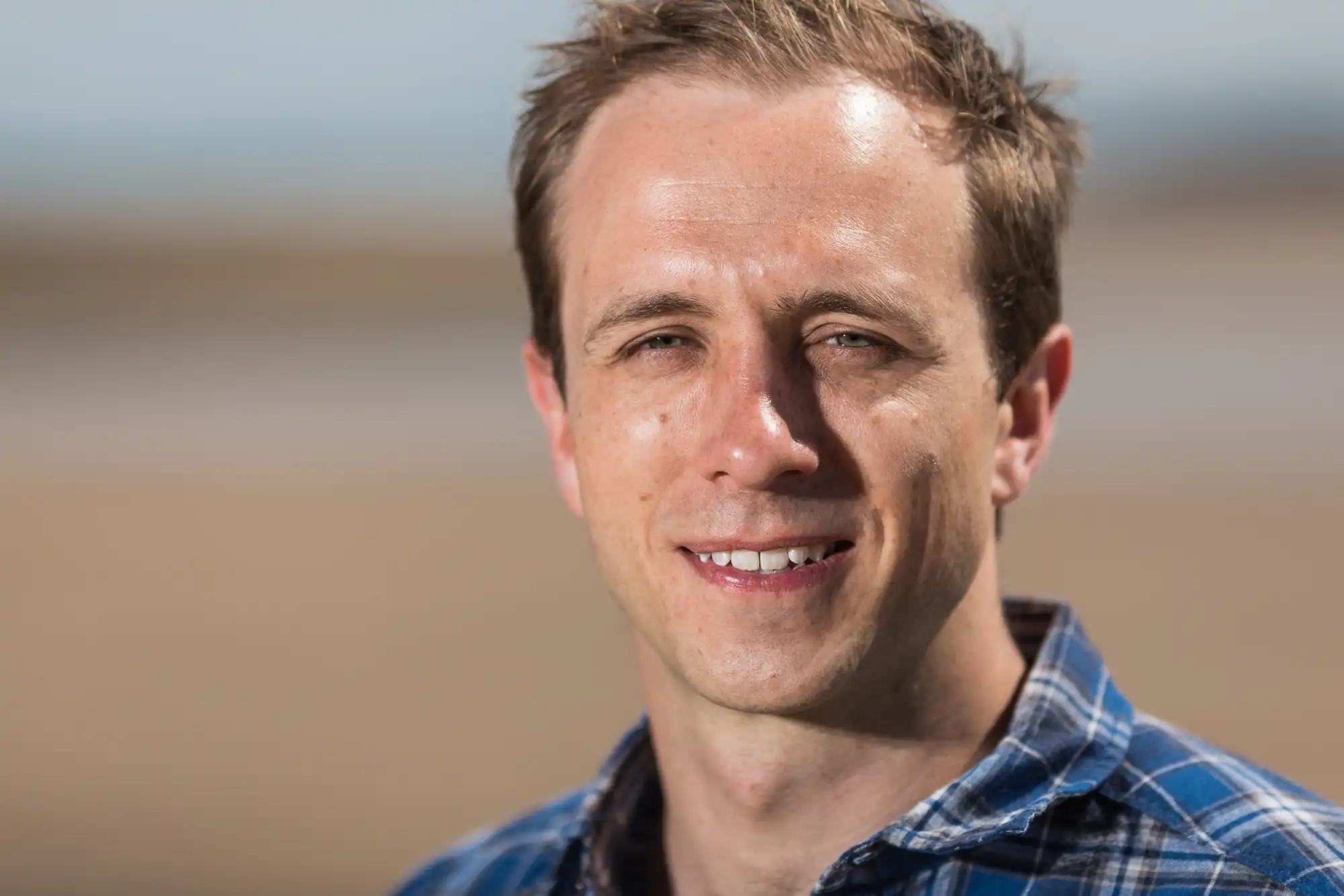A smiling man in a plaid shirt standing on a sandy beach with a blurred background.