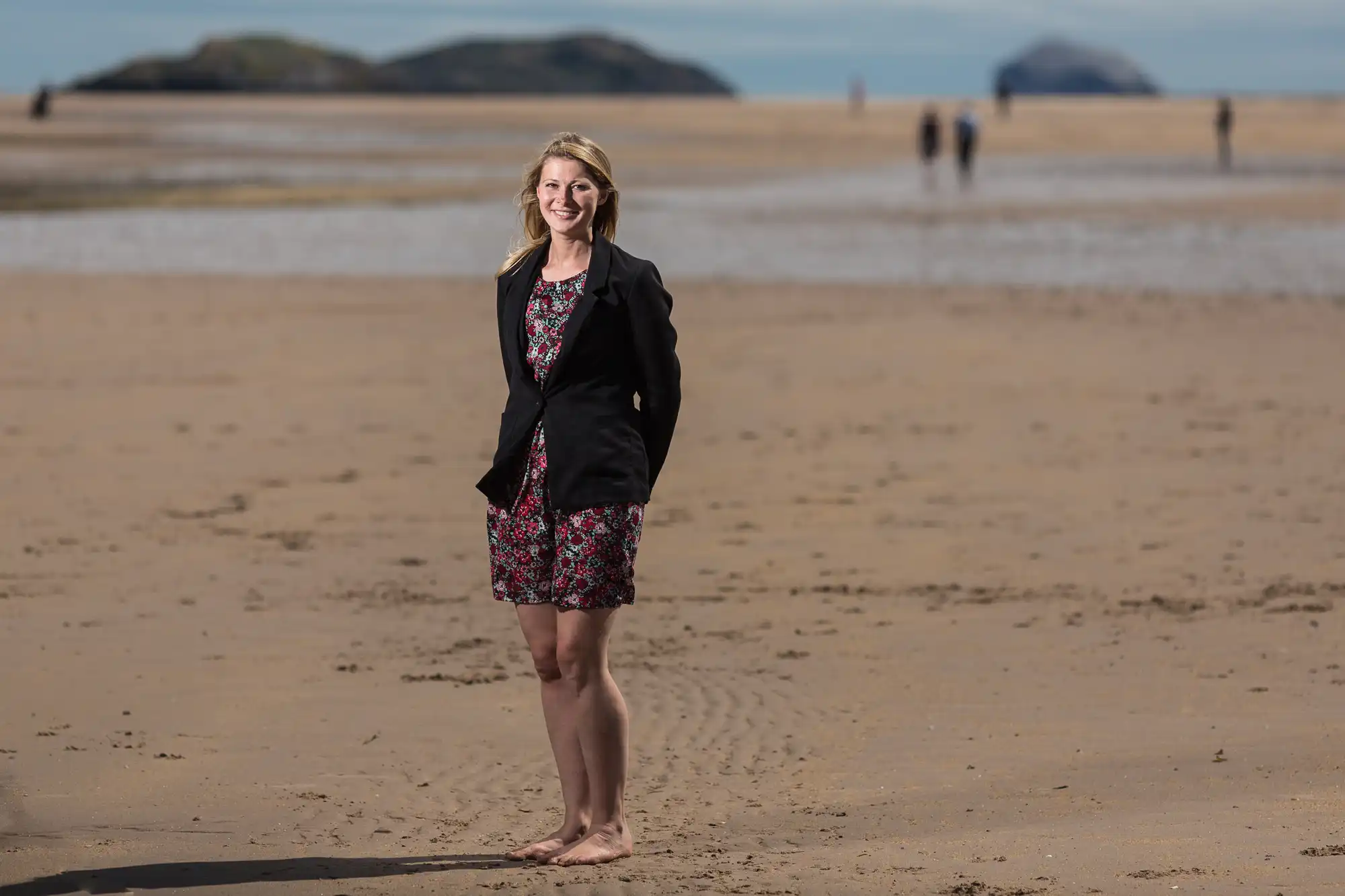 A woman in a floral dress and blazer standing barefoot on a sandy beach, smiling, with distant figures and rocks behind her.