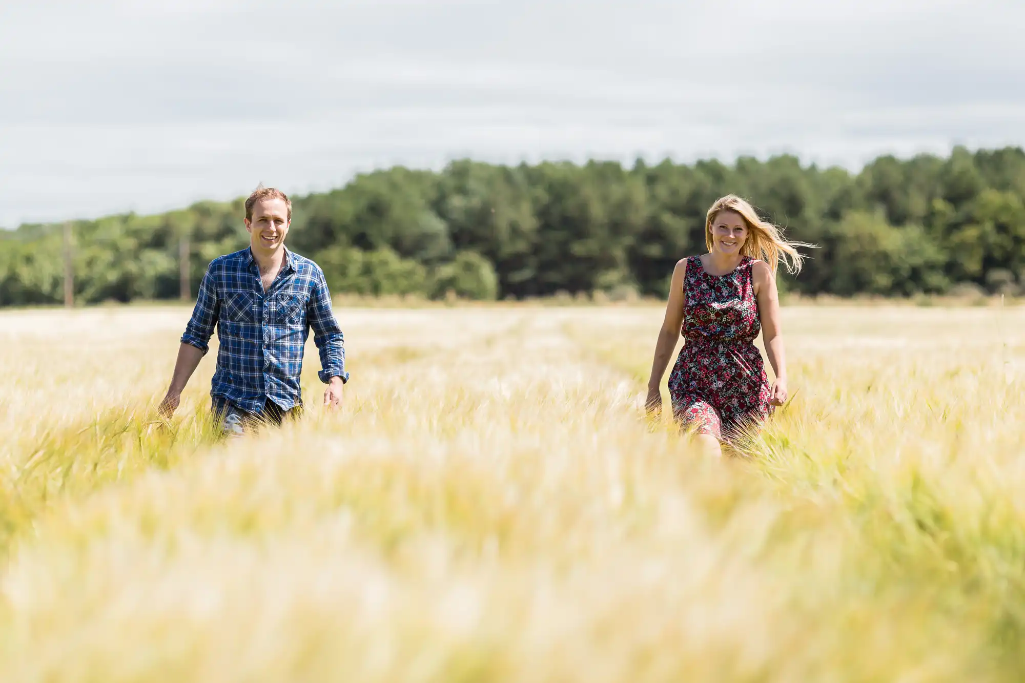 A man and a woman smiling and walking through a sunny wheat field, the man in a blue plaid shirt and the woman in a floral dress.