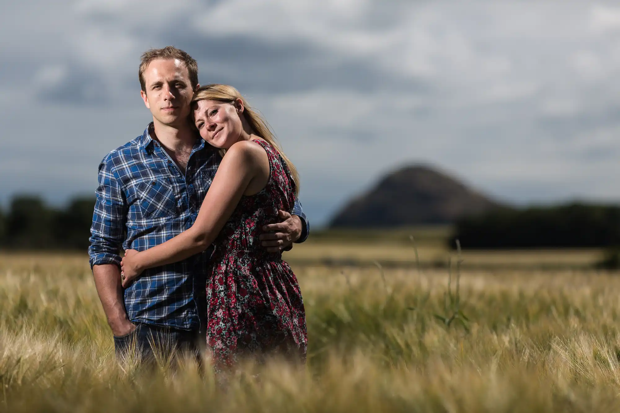 A couple embracing in a field with a cloudy sky above and a hill in the background.