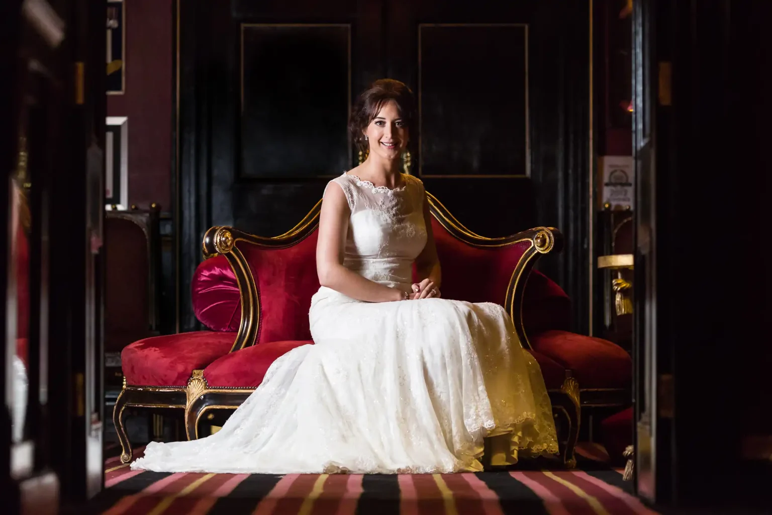 Luxury wedding photos at Prestonfield House Edinburgh - dramatic storytelling photography. A bride in a white lace wedding dress is seated on a red velvet sofa, smiling at the camera. The room has dark walls and ornate furnishings.