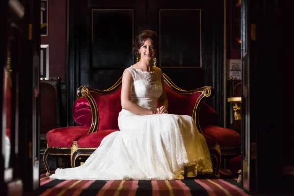 Luxury wedding photos at Prestonfield House Edinburgh - dramatic storytelling photography. A bride in a white lace wedding dress is seated on a red velvet sofa, smiling at the camera. The room has dark walls and ornate furnishings.