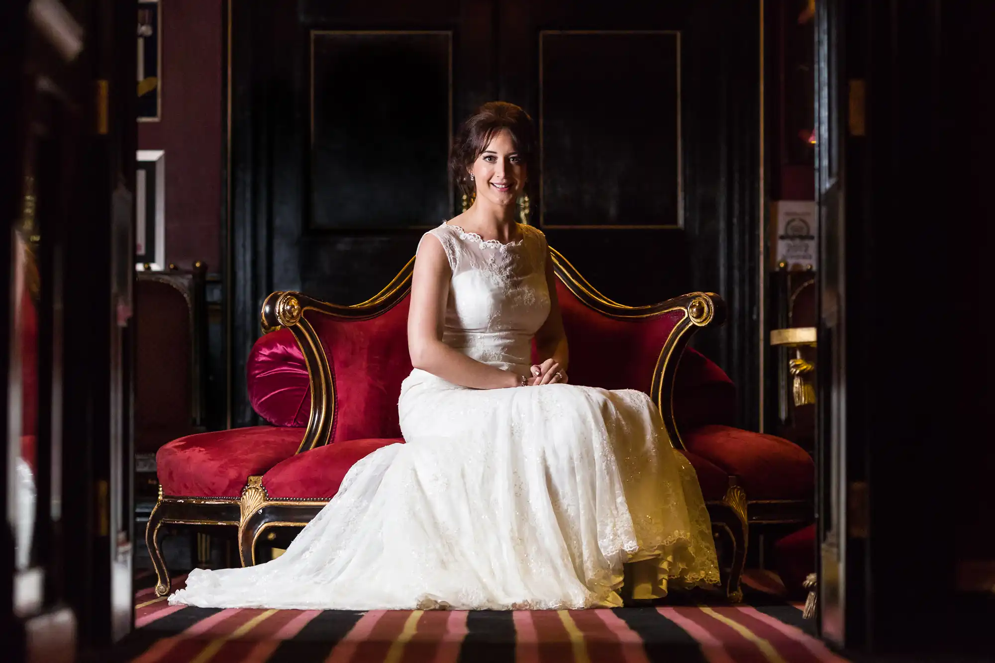 wedding photographer questions - bride sitting at Prestonfield House. A bride in a white lace wedding dress is seated on a red velvet sofa, smiling at the camera. The room has dark walls and ornate furnishings.