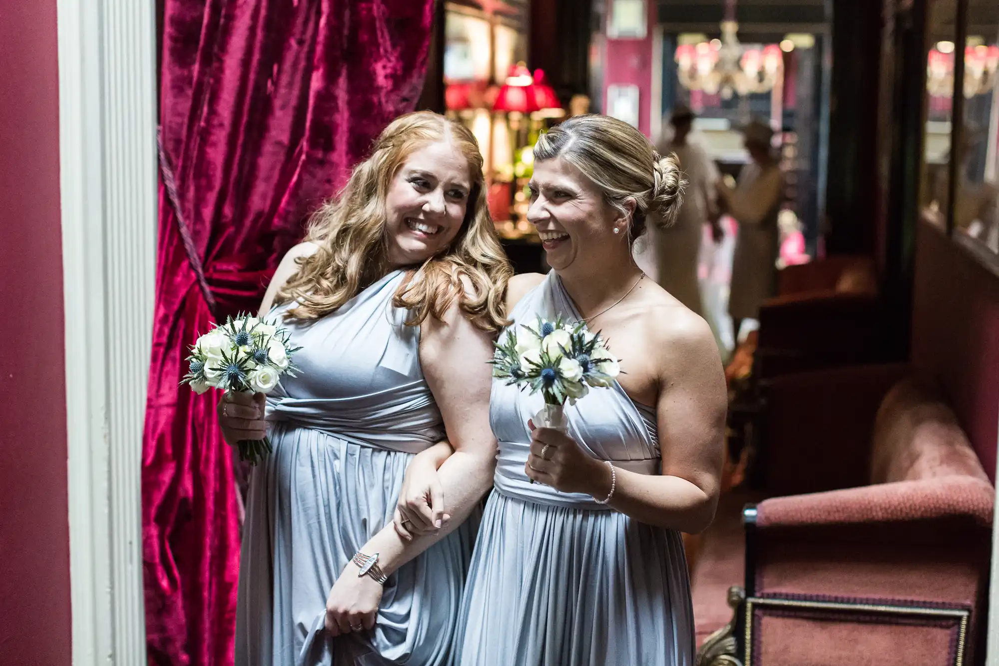 Two bridesmaids in lavender dresses hold bouquets and smile while walking arm-in-arm down a dimly lit corridor with red curtains and chairs.