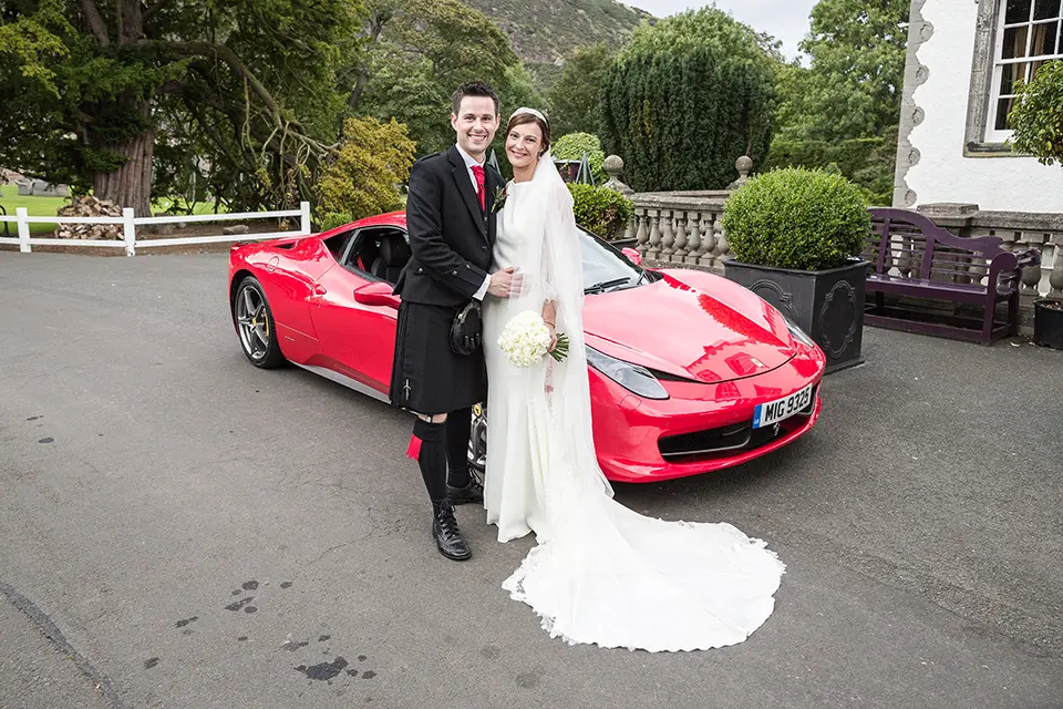 A bride and groom smiling beside a red sports car outside a scenic venue. the groom wears a kilt, and the bride is in a long white gown with a bouquet.
