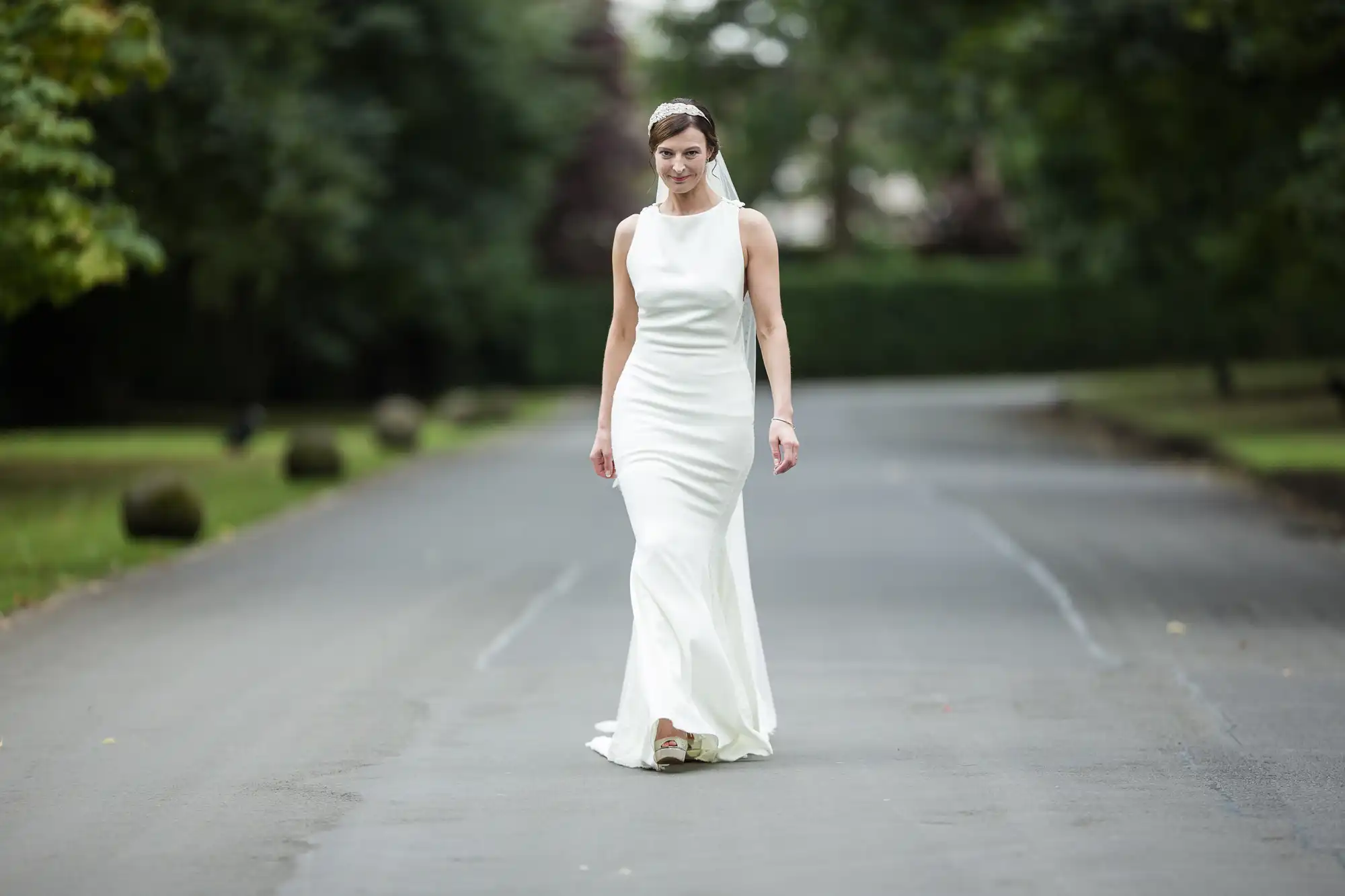 A bride in a sleeveless white dress is walking down a paved path surrounded by greenery.