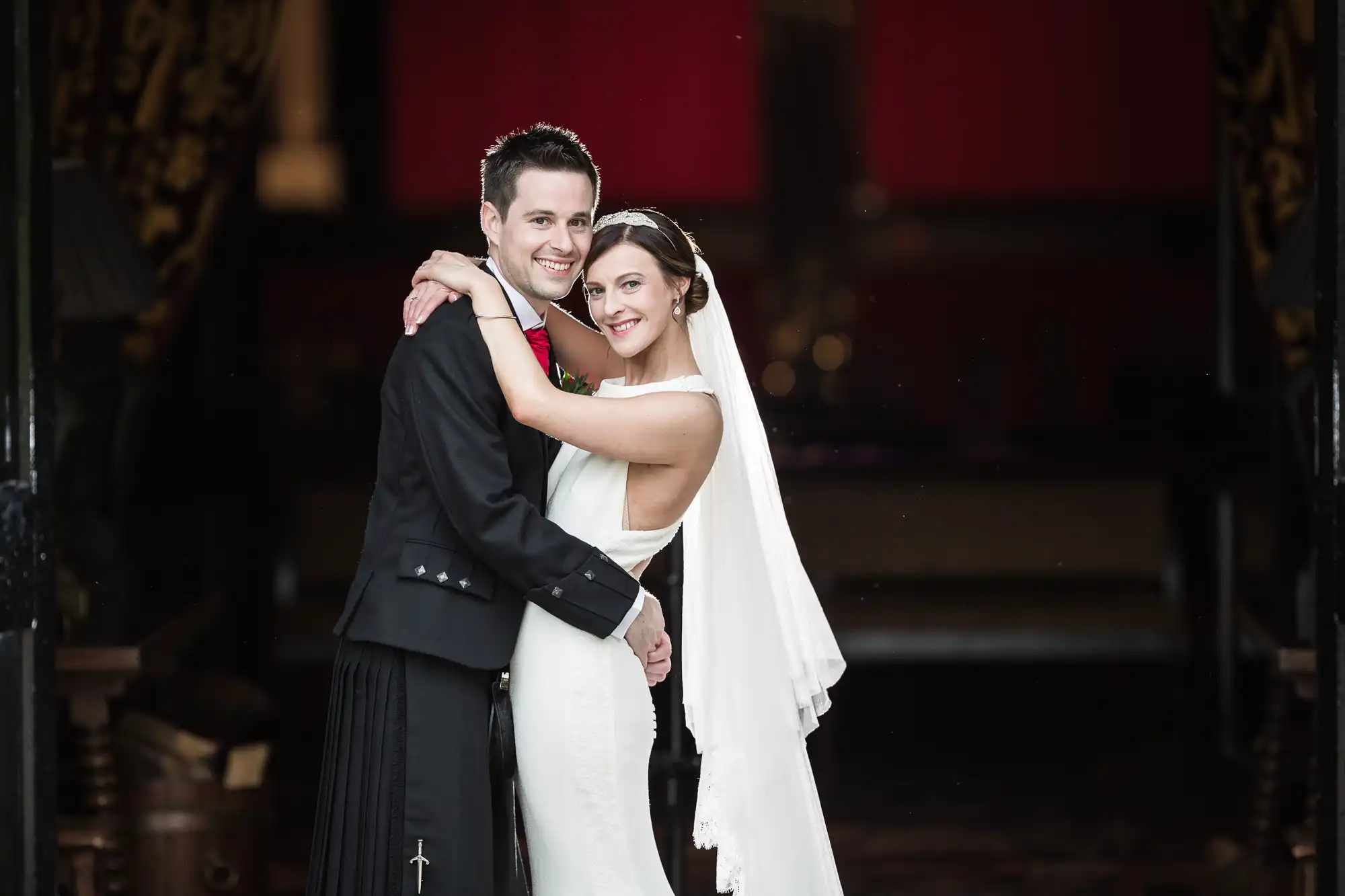 A bride in a white dress and veil stands with her arms around a groom in a black suit with a kilt. They are smiling and posing together indoors.