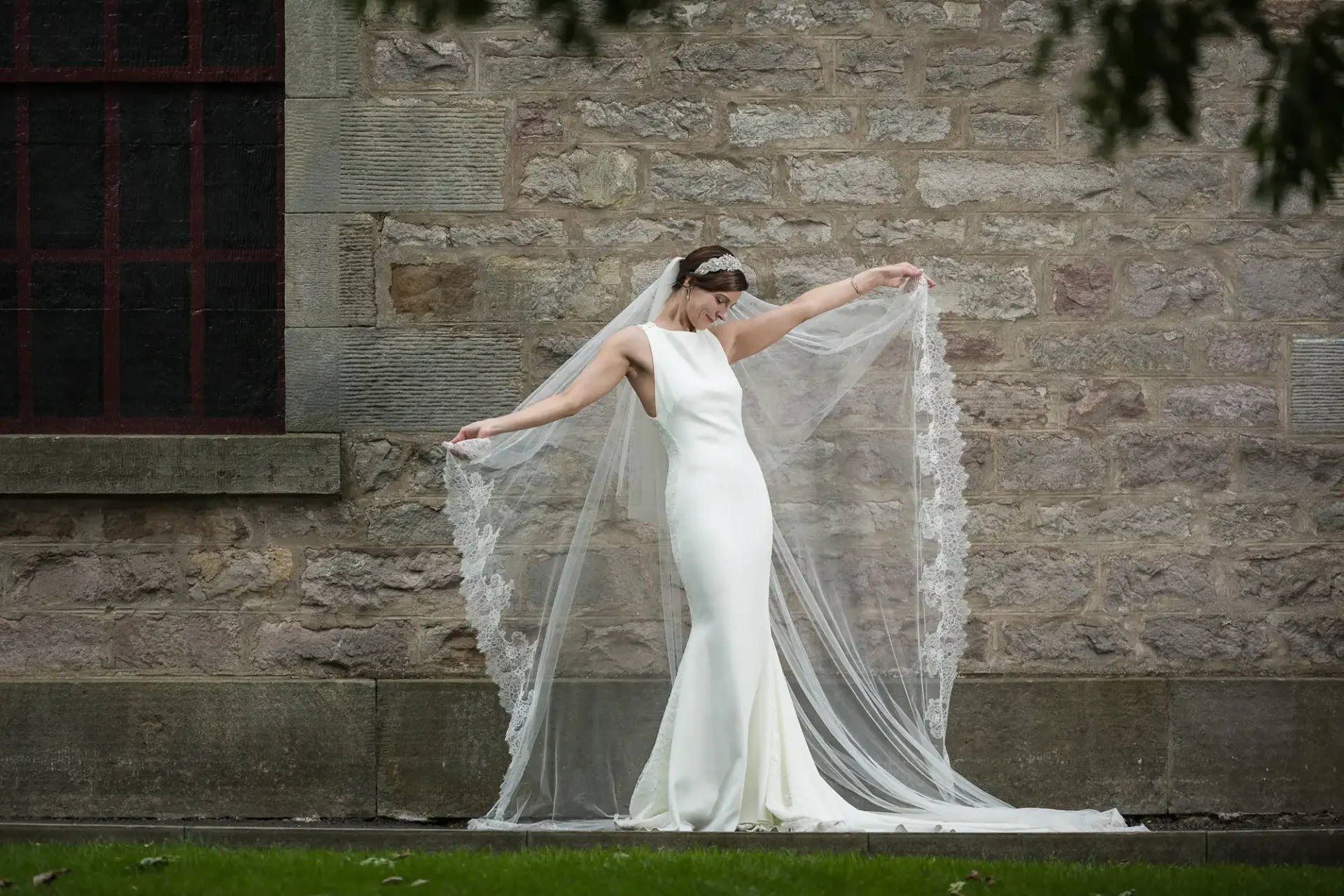 A bride in a sleeveless white wedding dress poses outside, holding out her lace-trimmed veil in front of a stone wall.