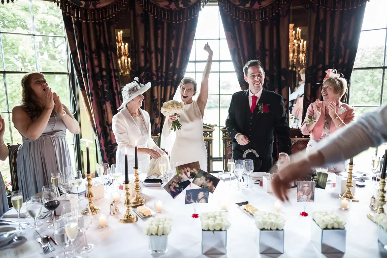 A bride in a white dress raises her arm in celebration at a wedding reception, surrounded by guests applauding and smiling. The table is decorated with flowers, candles, and framed photos.
