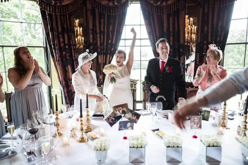 A bride in a white dress raises her arm in celebration at a wedding reception, surrounded by guests applauding and smiling. The table is decorated with flowers, candles, and framed photos.