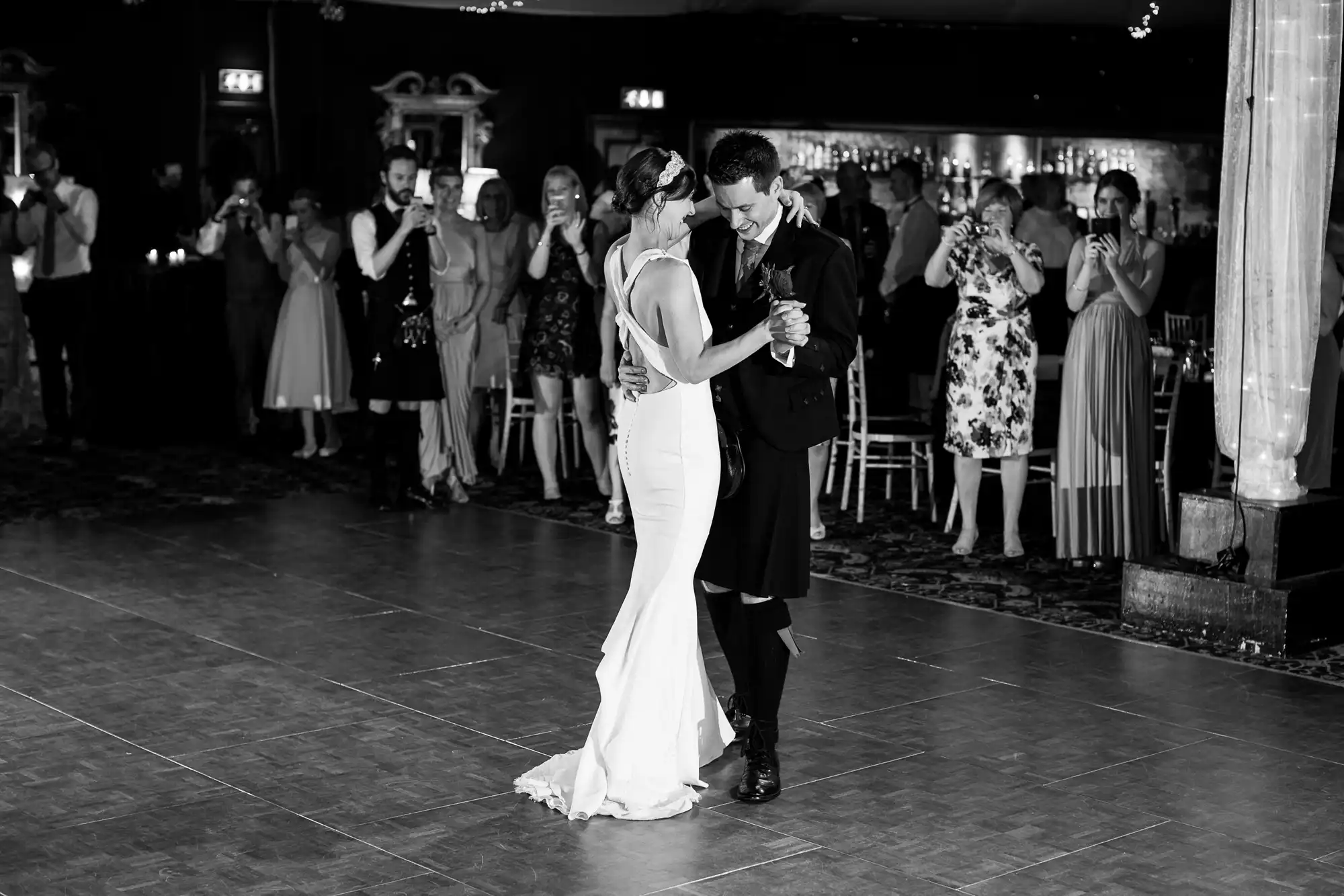 A couple dances closely together in the center of a ballroom while guests stand around them, taking photos and watching. The photo is in black and white.