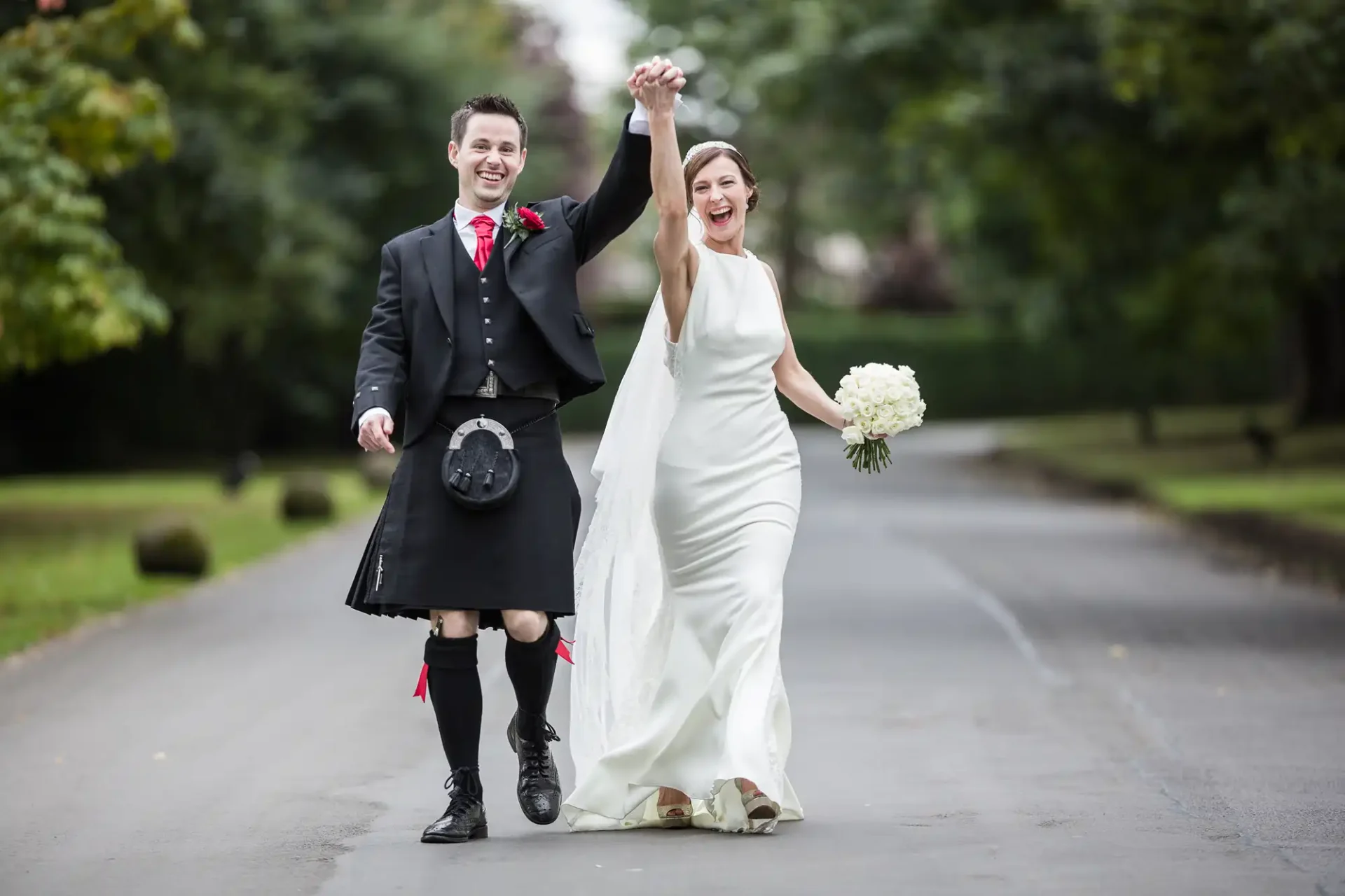A joyful couple, with the groom in a kilt and the bride in a white gown, walk hand-in-hand down a tree-lined path, celebrating their wedding day at Prestonfield House - Jen and David