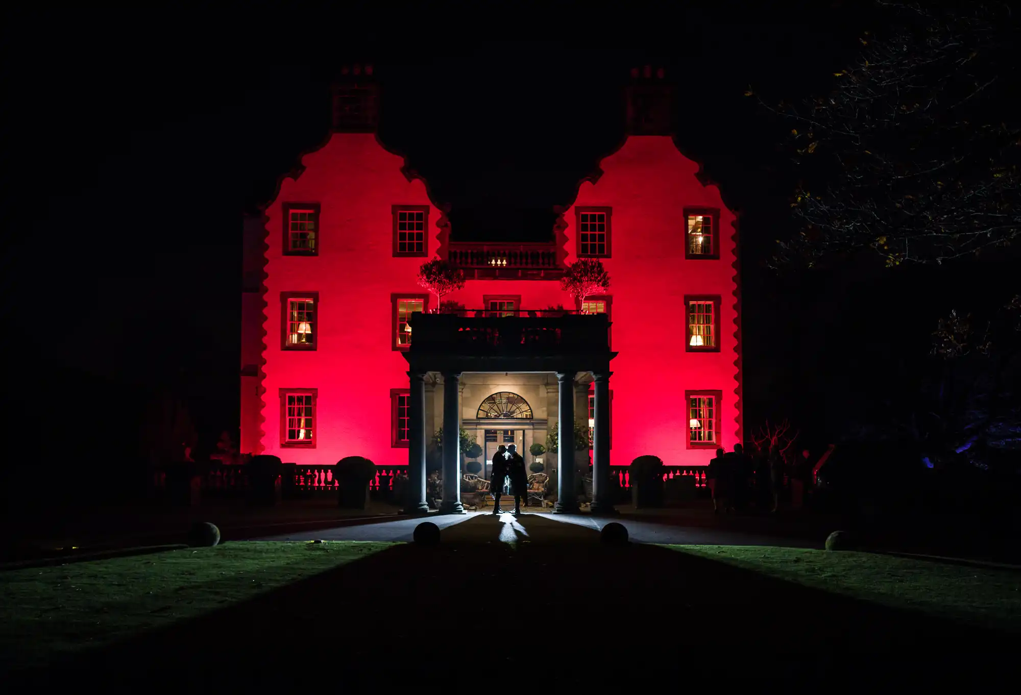 A newlywed couple stands in front of a large building with red-lit walls and illuminated windows at night.