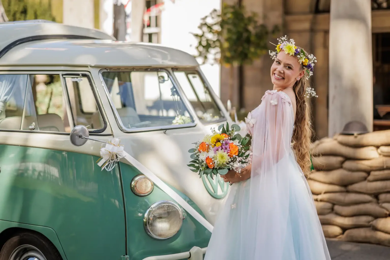 A woman in a white dress with floral headpiece holds a bouquet of flowers and stands beside a vintage green and white van decorated with ribbons.