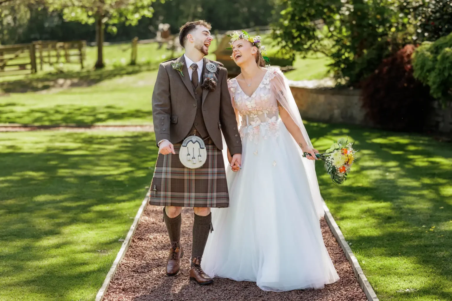 A bride in a white dress and a groom in a kilt are holding hands and smiling at each other outdoors on a sunny day.