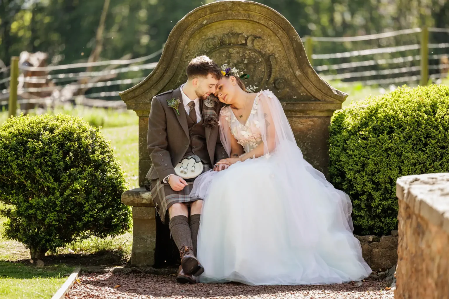 A bride and groom sit closely together on a stone bench outdoors, with the bride resting her head on the groom’s shoulder. Both are wearing wedding attire.