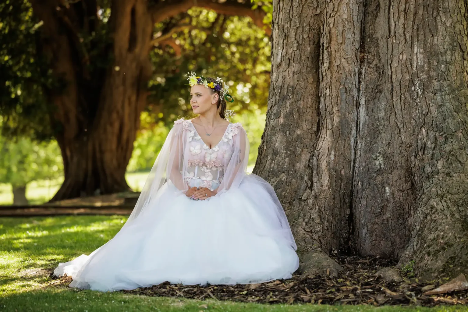 A woman wearing a white wedding dress and flower crown sits on the grass, leaning against a large tree trunk in a sunlit park.