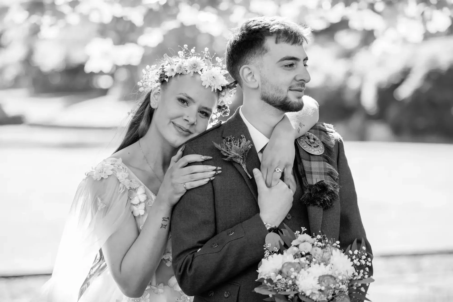 A bride rests her head on the groom’s shoulder as they pose outdoors; she smiles gently while he looks ahead, holding a bouquet.