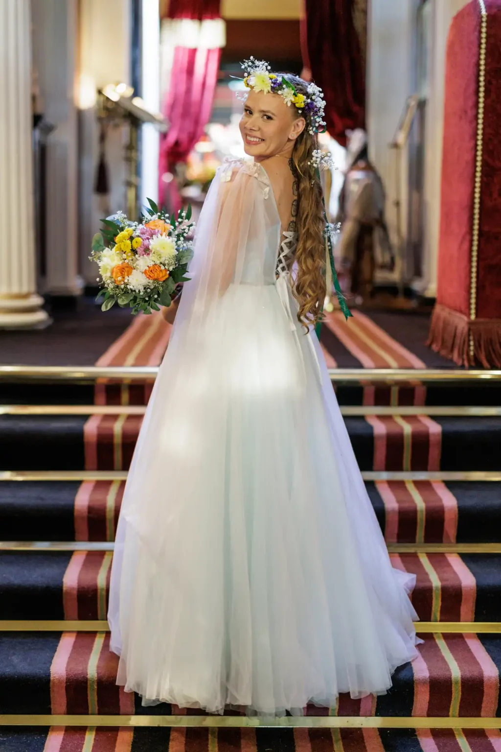 A woman in a white wedding dress and flower crown stands on carpeted stairs, holding a colorful bouquet and smiling over her shoulder.