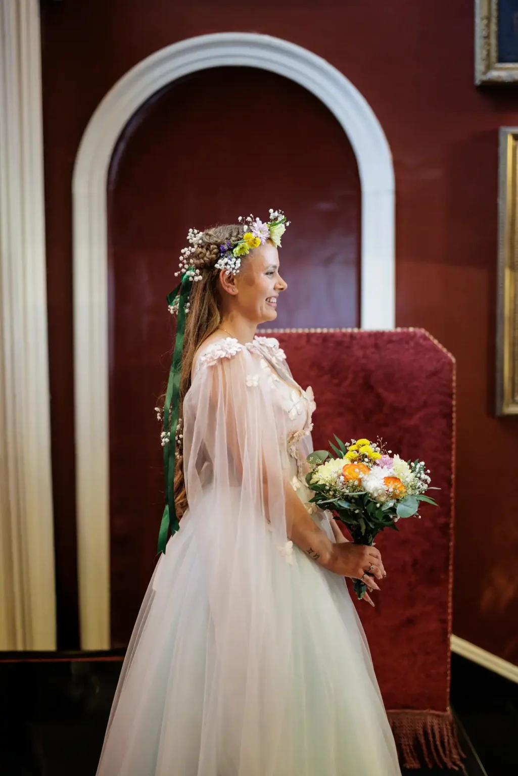 A woman in a white dress and flower crown holds a bouquet of flowers, standing indoors in front of a red background and white archway.