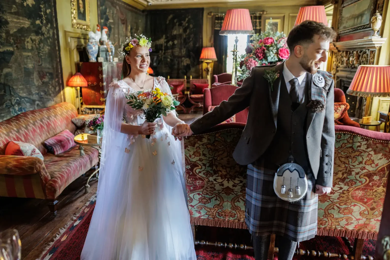 A bride in a white dress and floral crown holds a bouquet and the hand of a groom in a kilt and jacket inside an ornately decorated, vintage-style room.