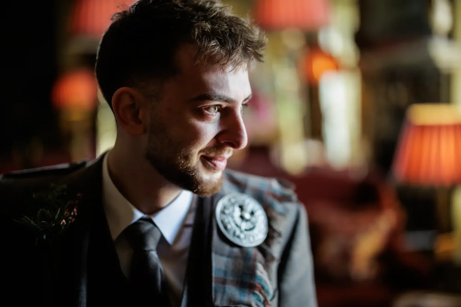 A man in a suit with a tartan sash and decorative brooch looks to the side in a warmly lit room with red lamps in the background.